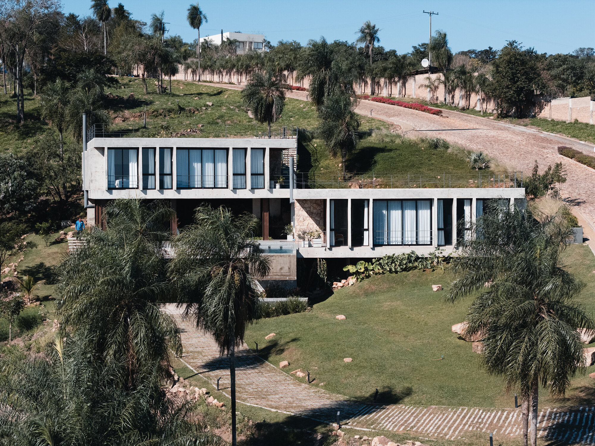 Tiered concrete residence on a sloped site with palms and native vegetation under a clear sky