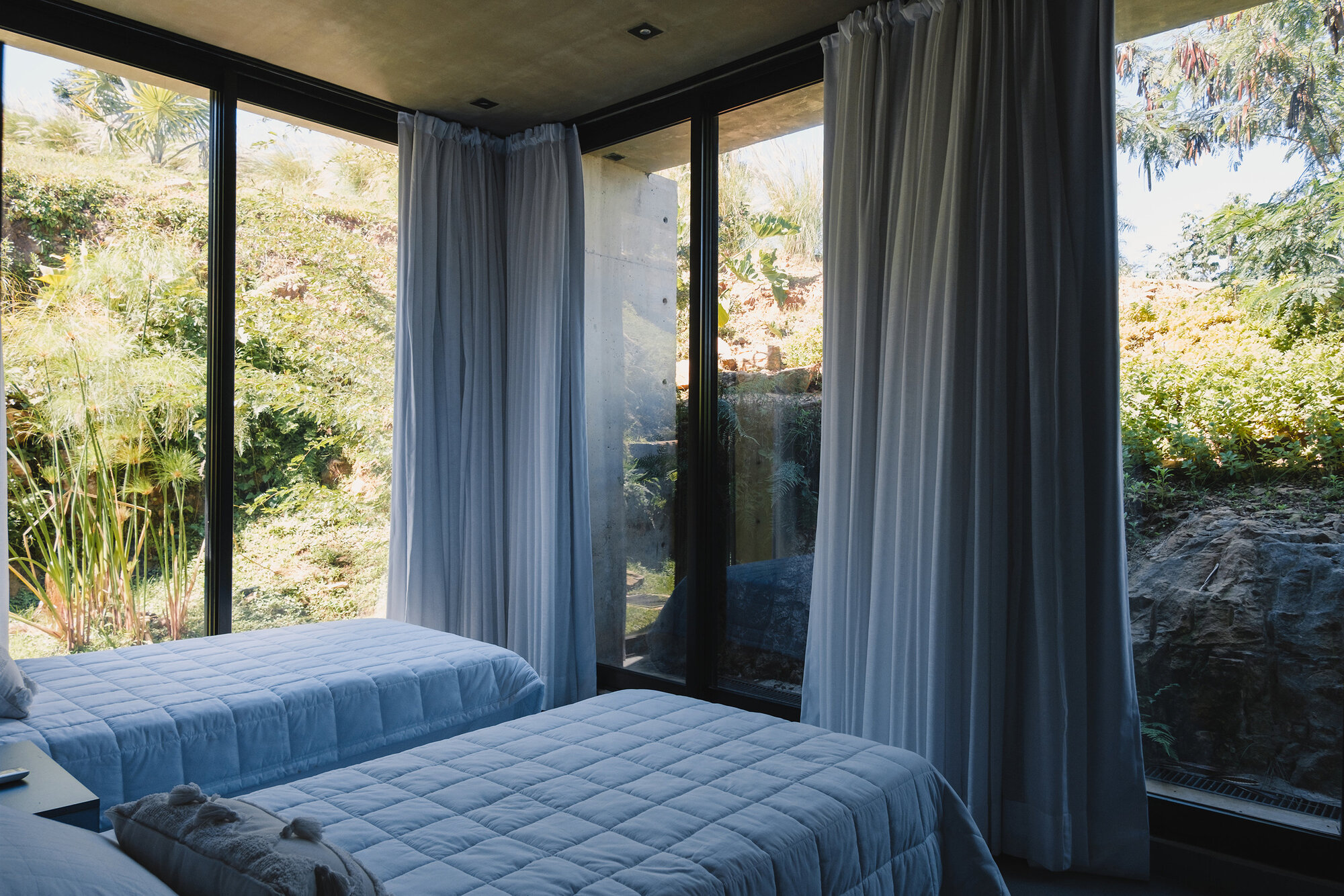 Corner bedroom with floor-to-ceiling glass walls, grey curtains, and twin beds facing vegetation