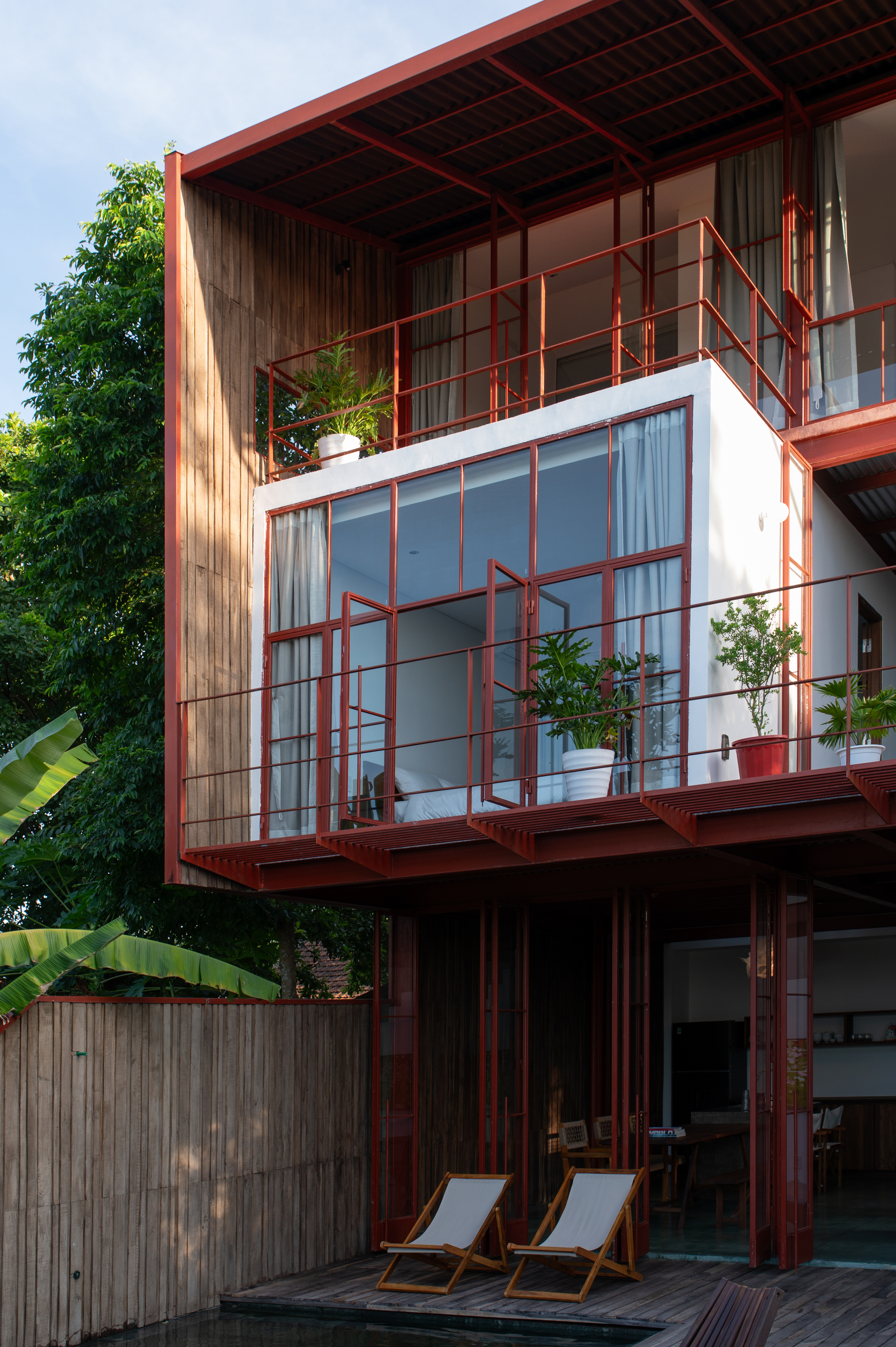 Stacked balconies with red metal railings and timber cladding framed by tropical foliage