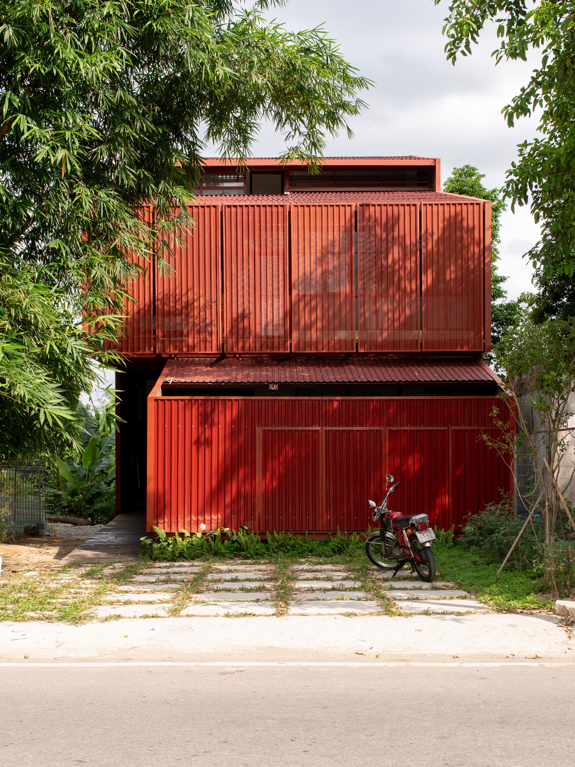 Street facade of red corrugated metal volumes with a motorcycle parked beside stepping stone path