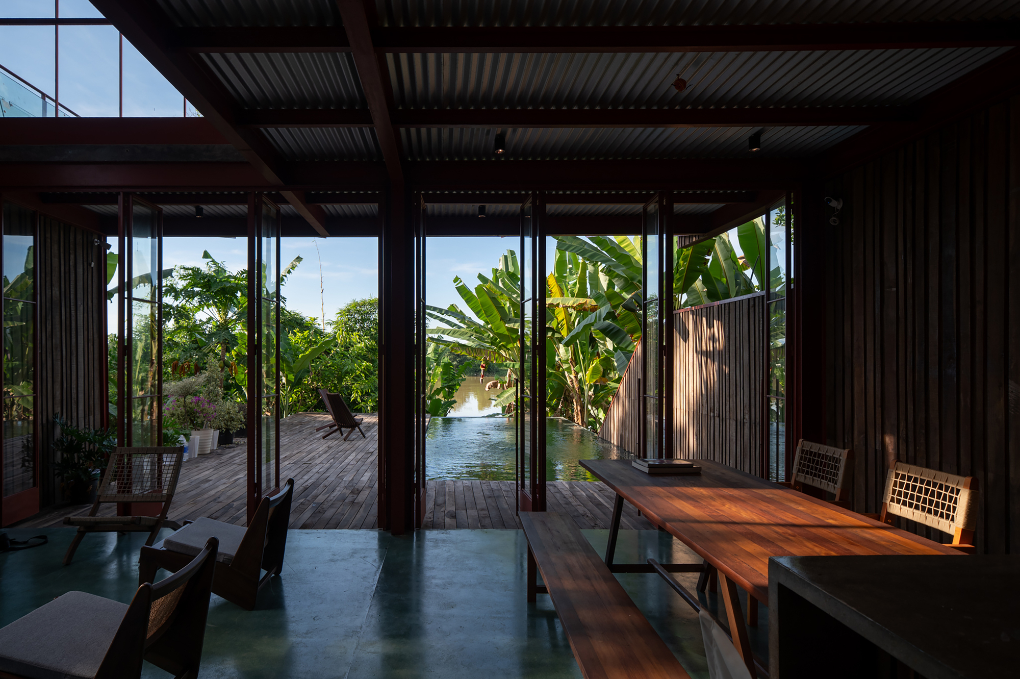 Interior view through open glass doors to timber deck and pond surrounded by banana palms at dusk