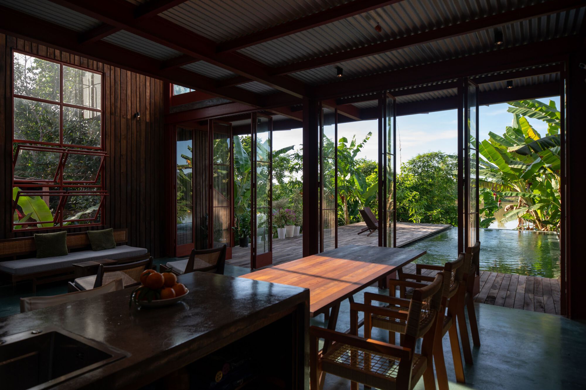 Open living pavilion with folding glass walls framing view of terrace pool and banana trees beyond