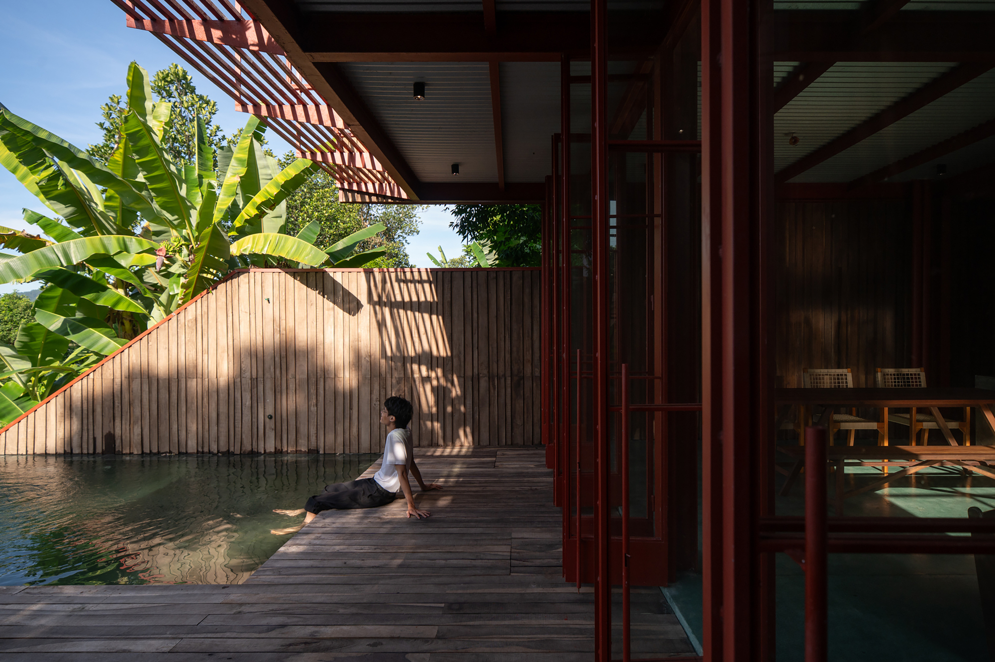 Covered timber deck alongside a reflecting pool with a figure seated in morning light