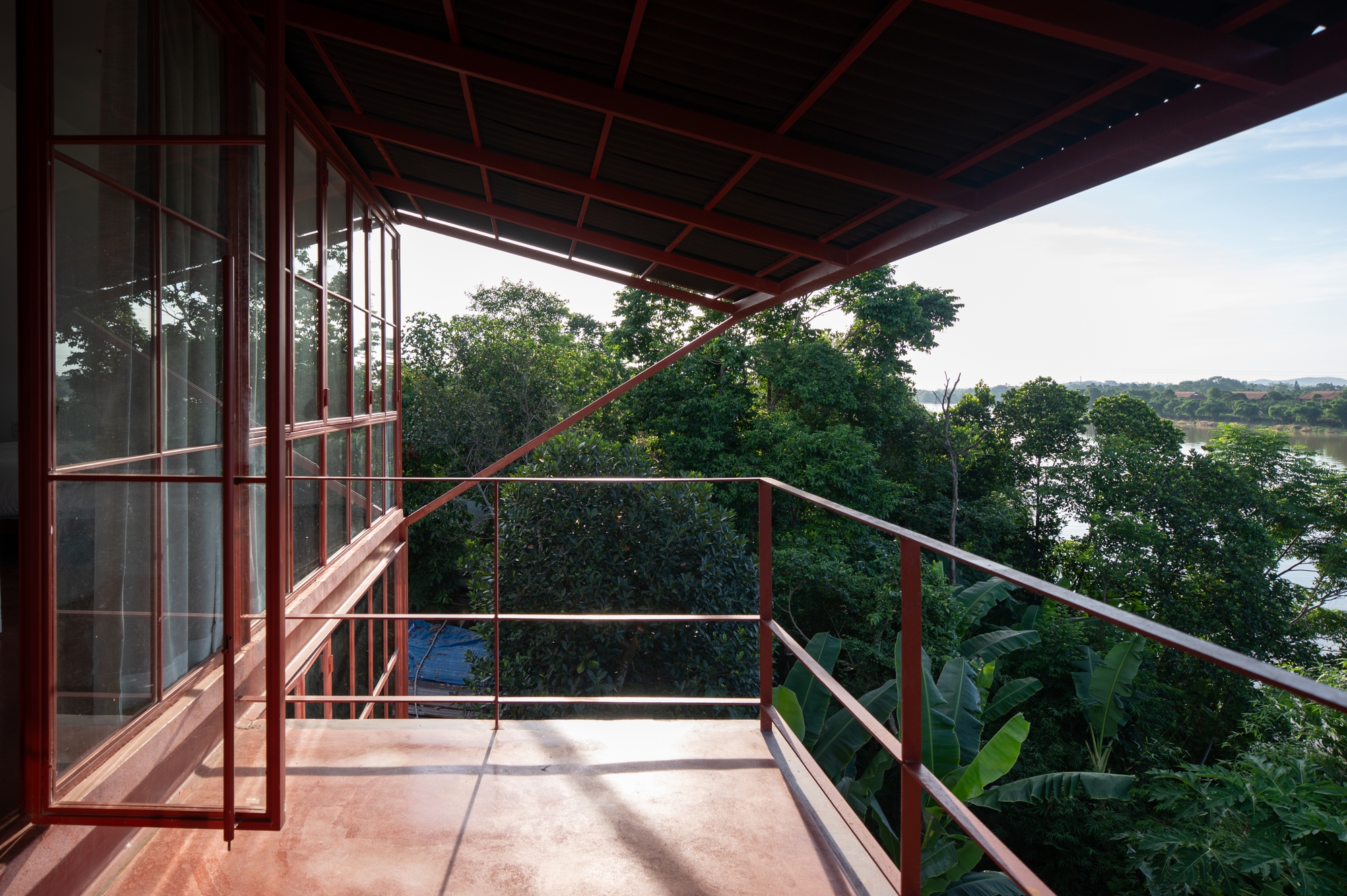 Upper-level terrace with red steel trellis overhead looking toward tree canopy and distant water