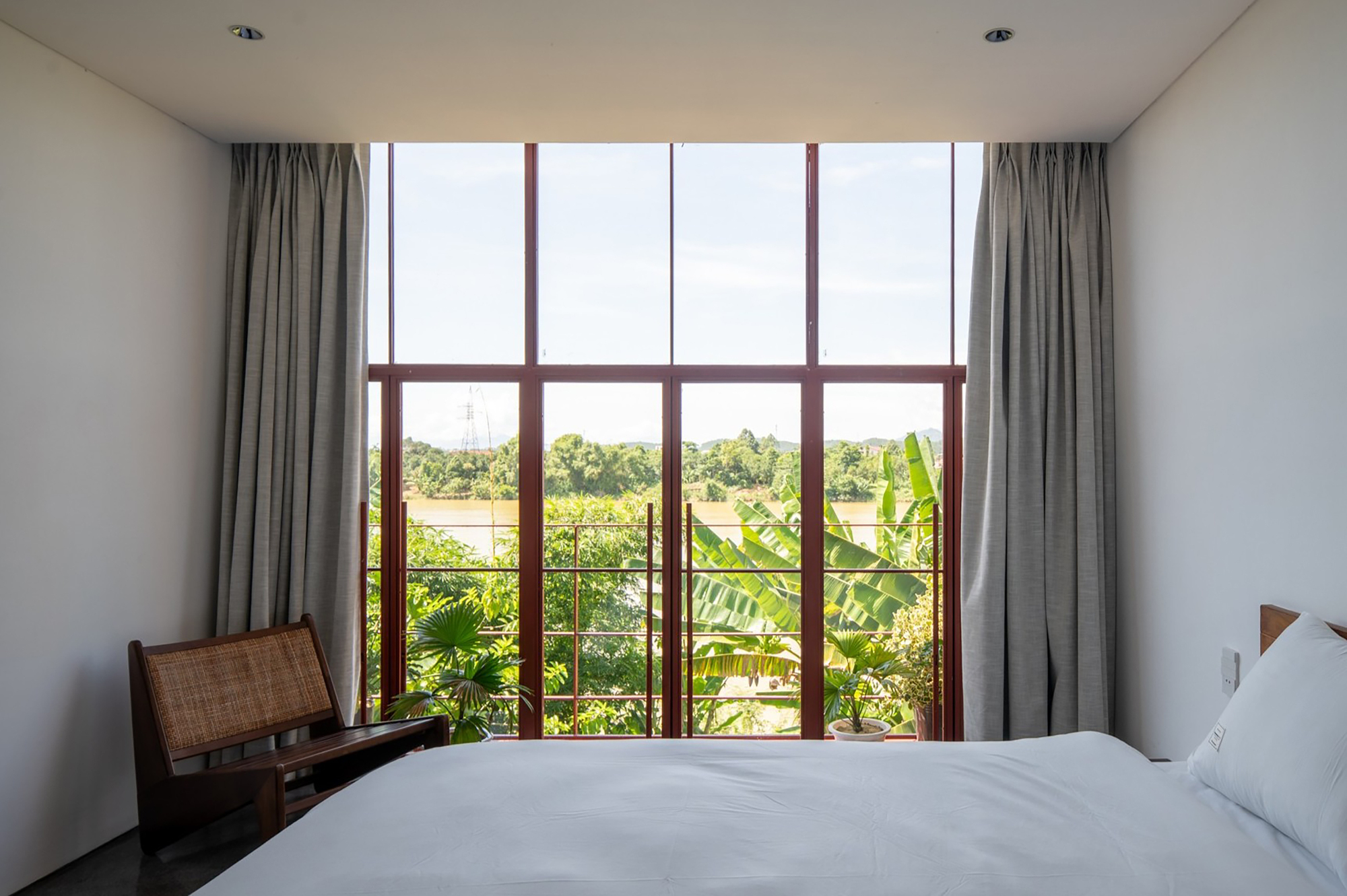 Bedroom interior with floor-to-ceiling red steel framed windows overlooking banana palms and river