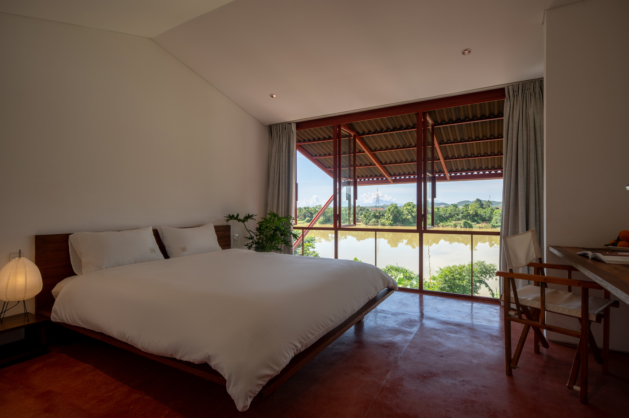 Bedroom with exposed steel truss roof and glazed doors opening to a river view beyond