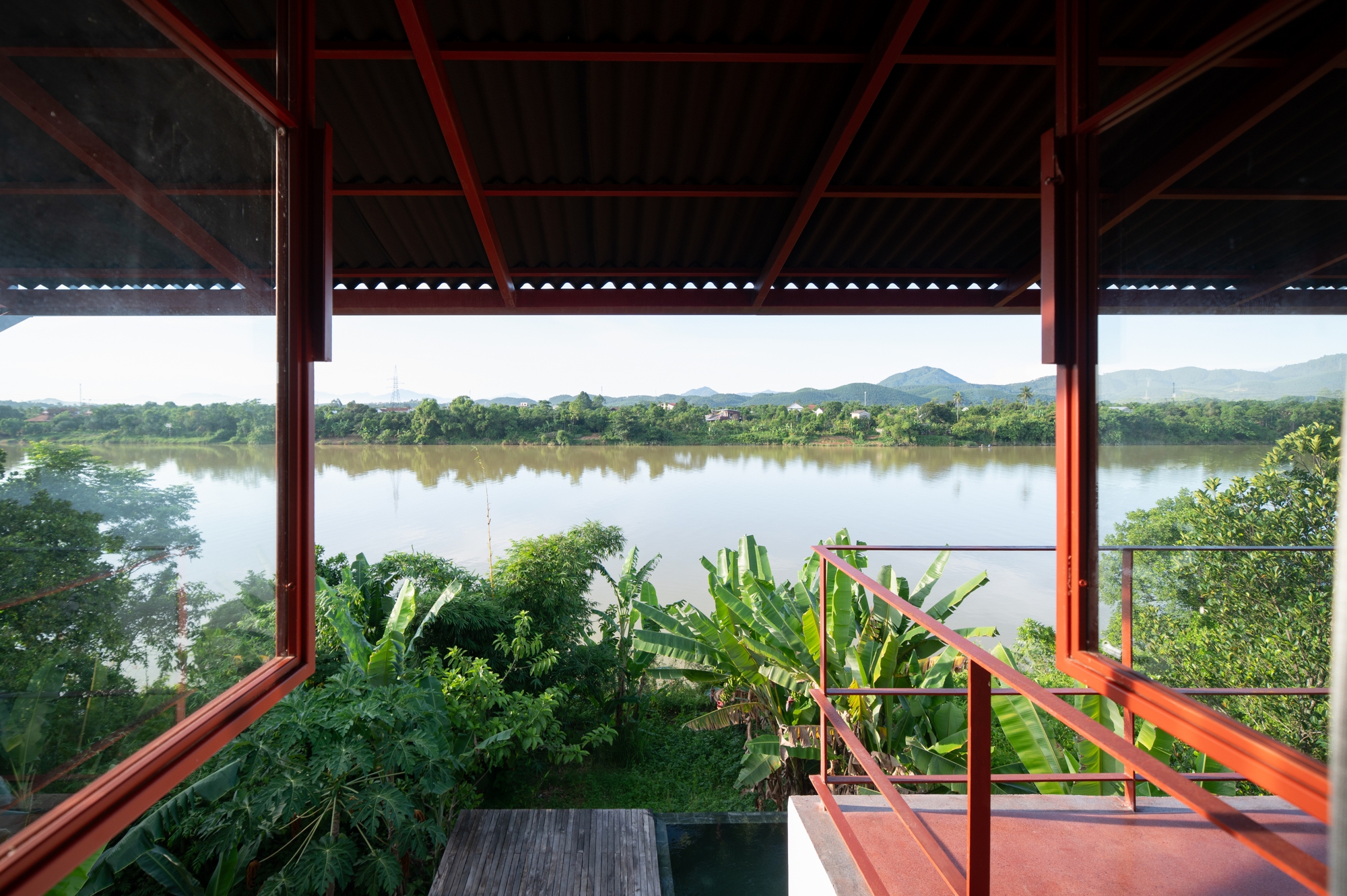 Covered terrace with red steel structural frame and glass railing facing the river and distant mountains