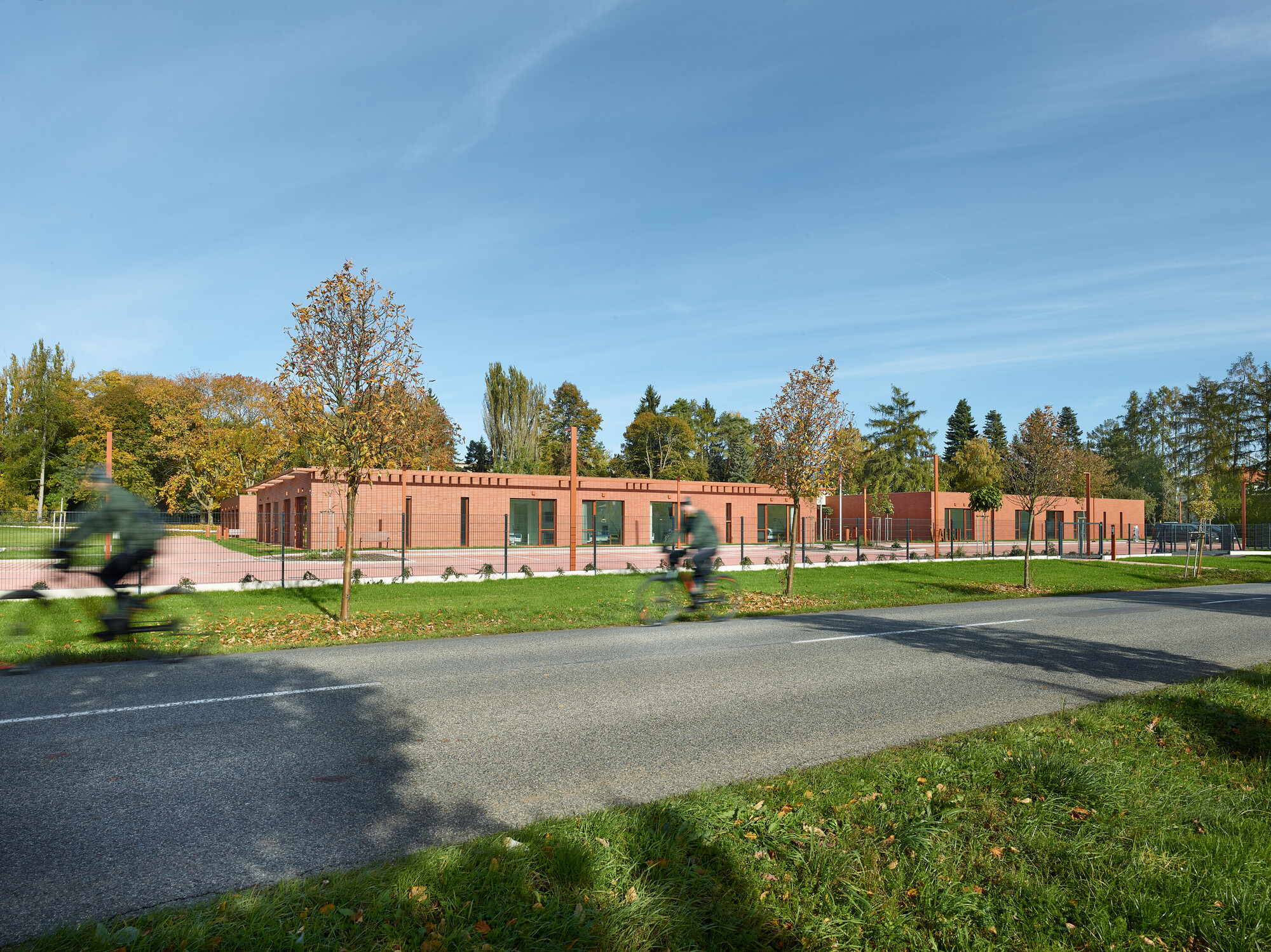 Long red brick facade with regularly spaced windows facing a park and road with cyclists in autumn