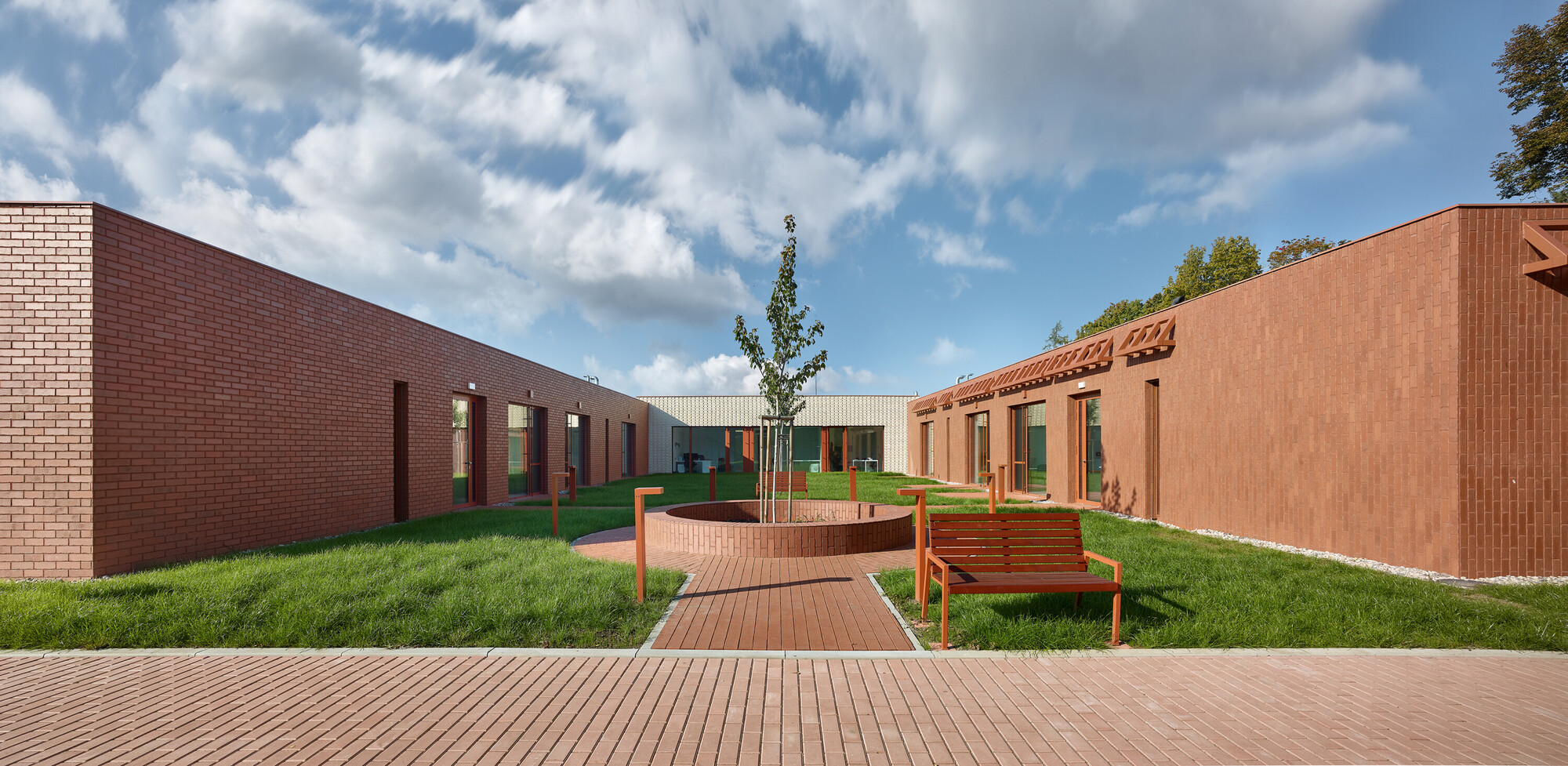 Courtyard with timber decking, circular planter and benches surrounded by red brick and tile walls under clouds