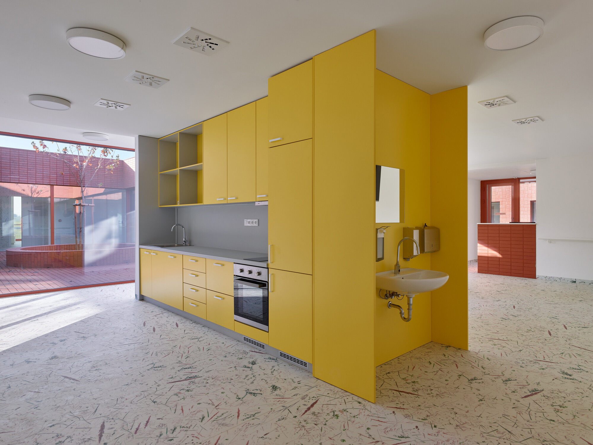 Yellow kitchen volume with integrated sink and terrazzo floor looking toward the courtyard through glazing