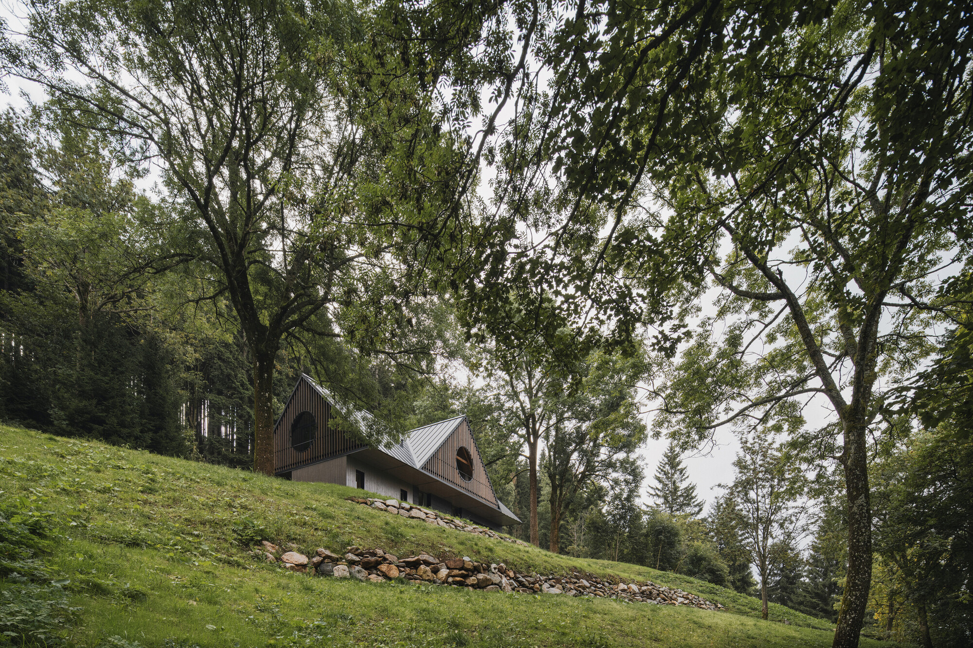 Side view of the dual gable volumes on a grassy hillside with stone retaining wall