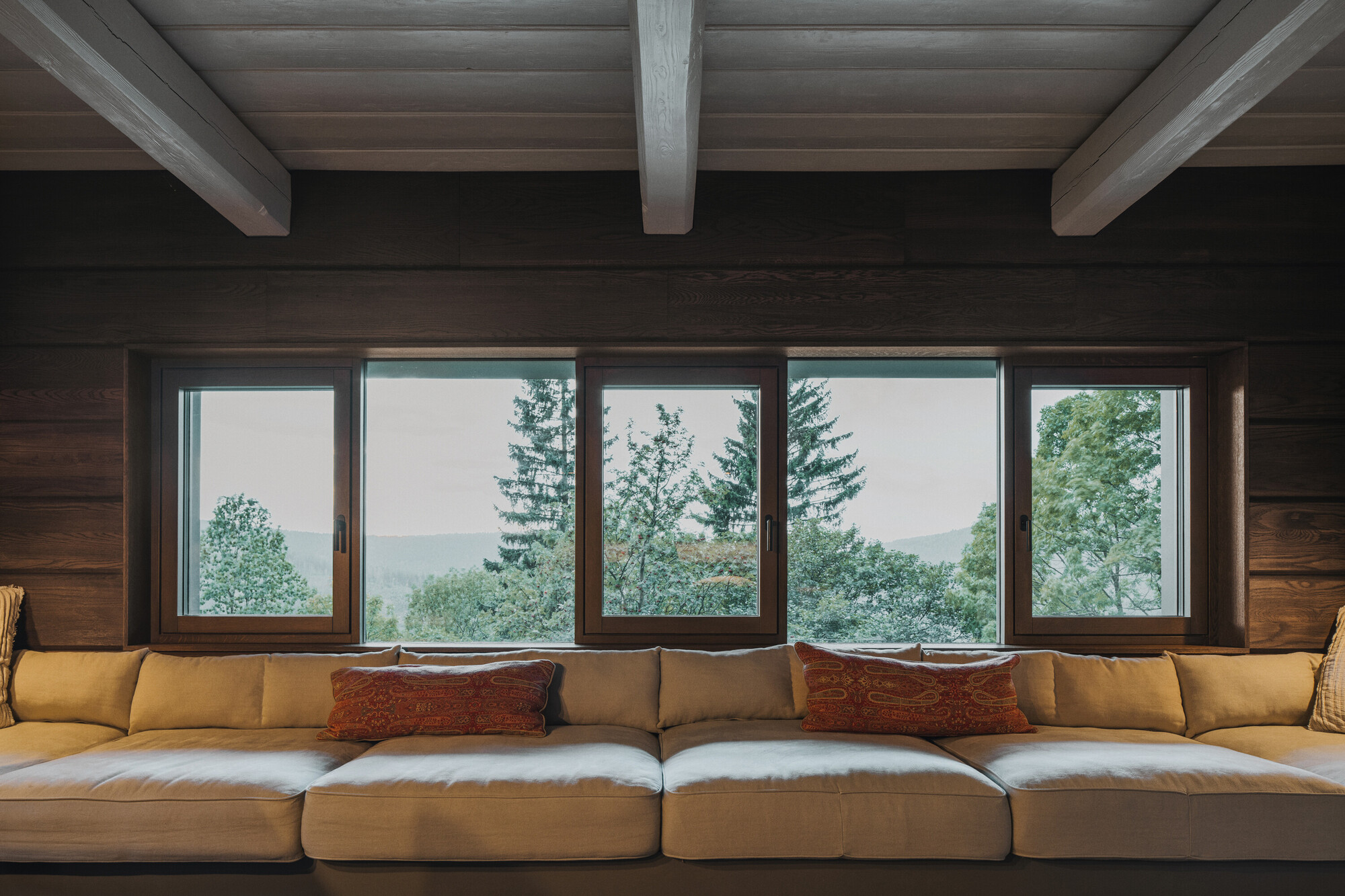 Five-panel window wall framing misty forest views above a seating area with exposed ceiling beams