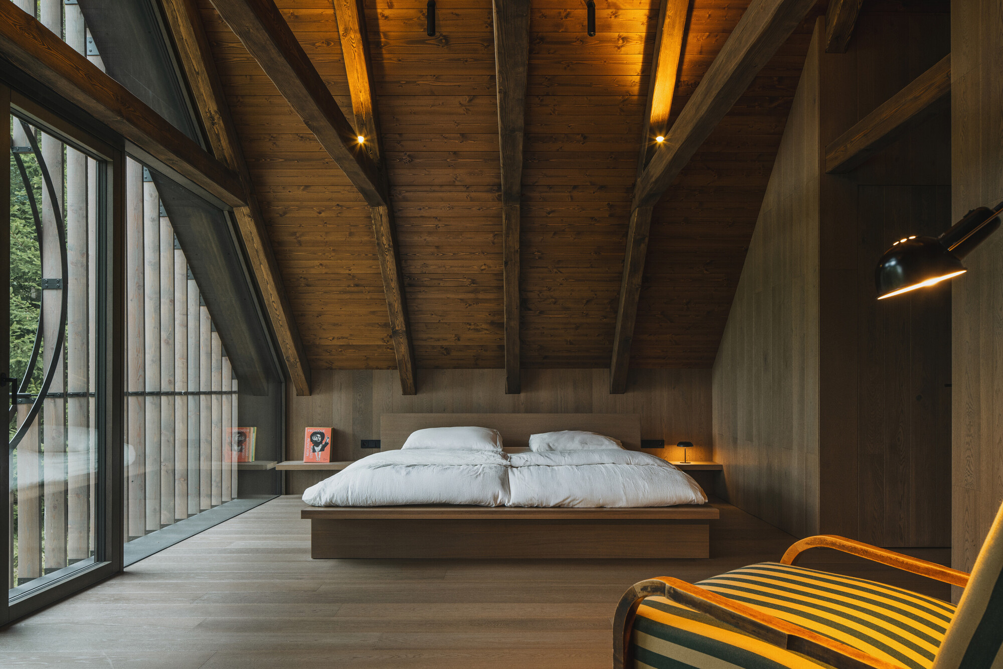 Bedroom beneath pitched timber roof with glazed wall overlooking trees at dusk