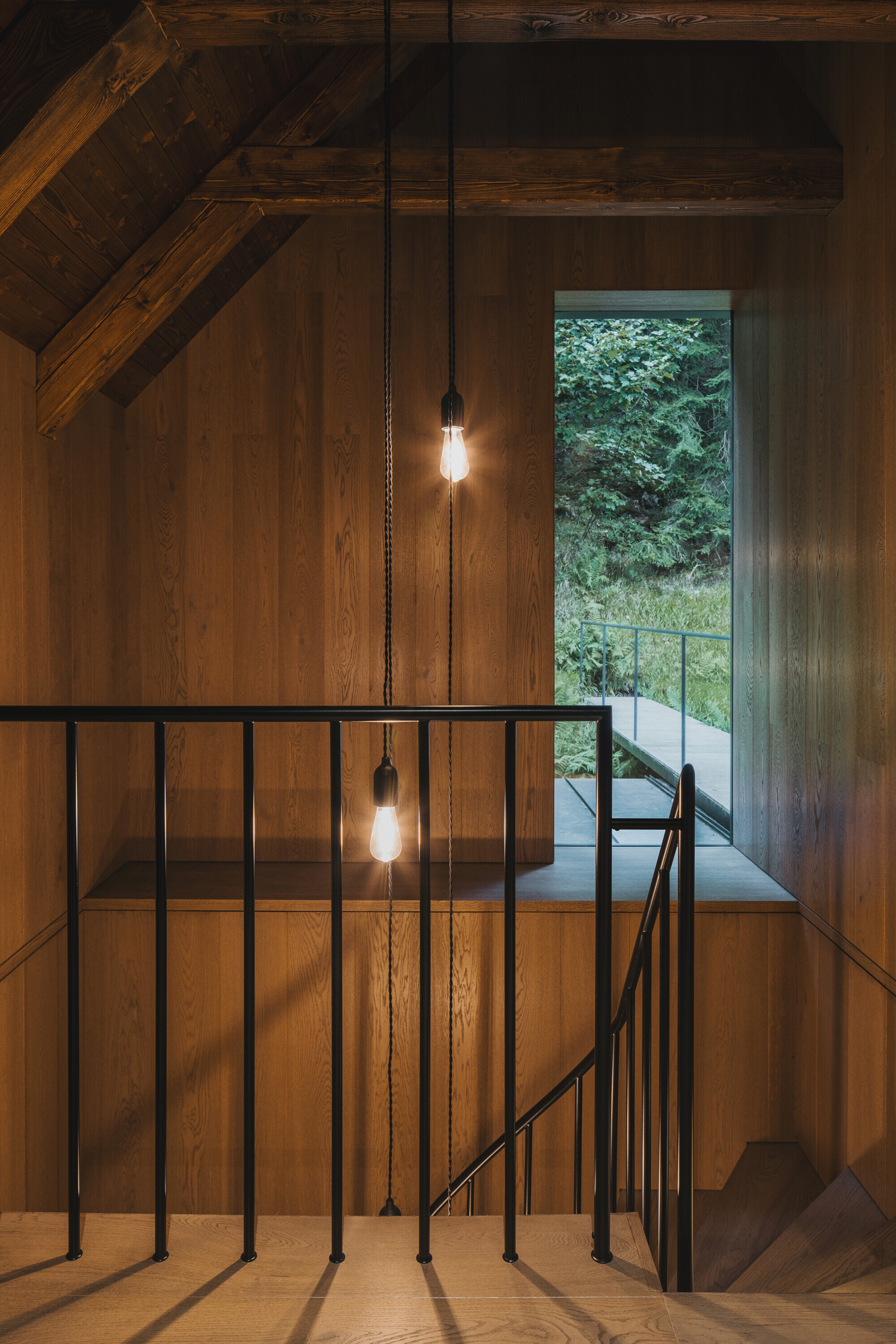 Timber-lined stairwell with black metal railing and pendant bulbs illuminated at dusk