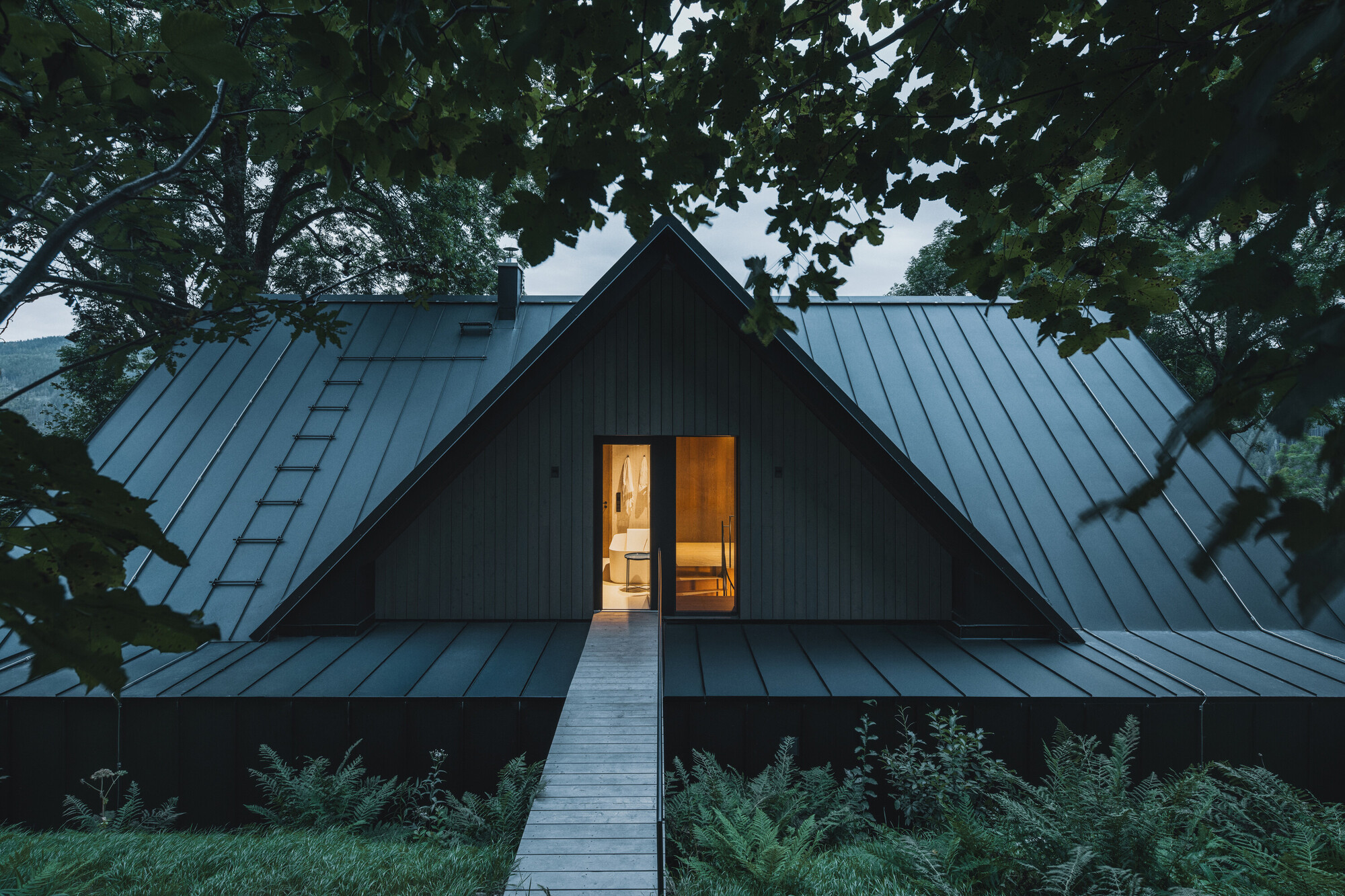 Gabled metal roof with central glazed gable illuminated at twilight among mature trees