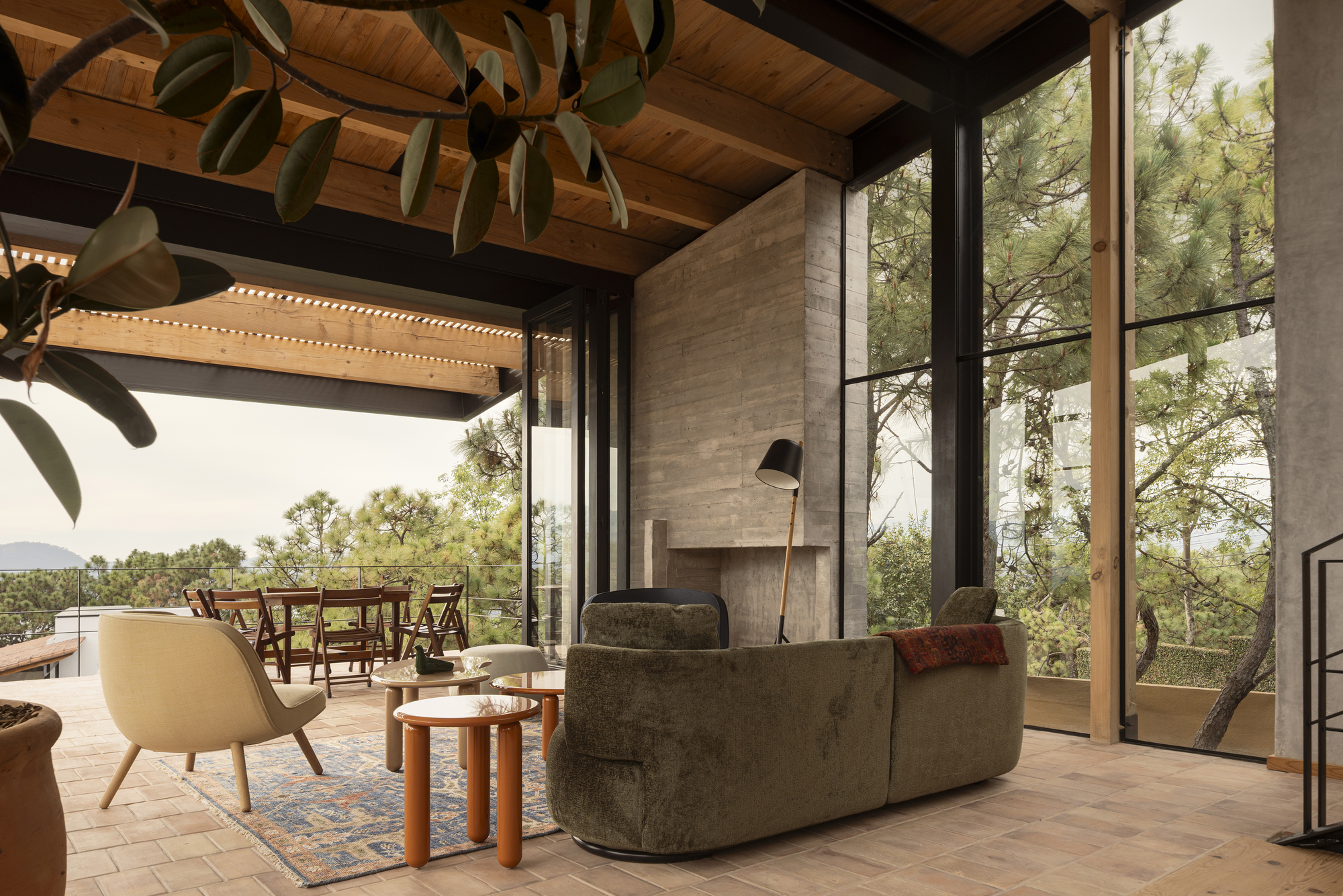 Living area opening to a terrace with exposed timber ceiling beams and steel frame glazing overlooking forest