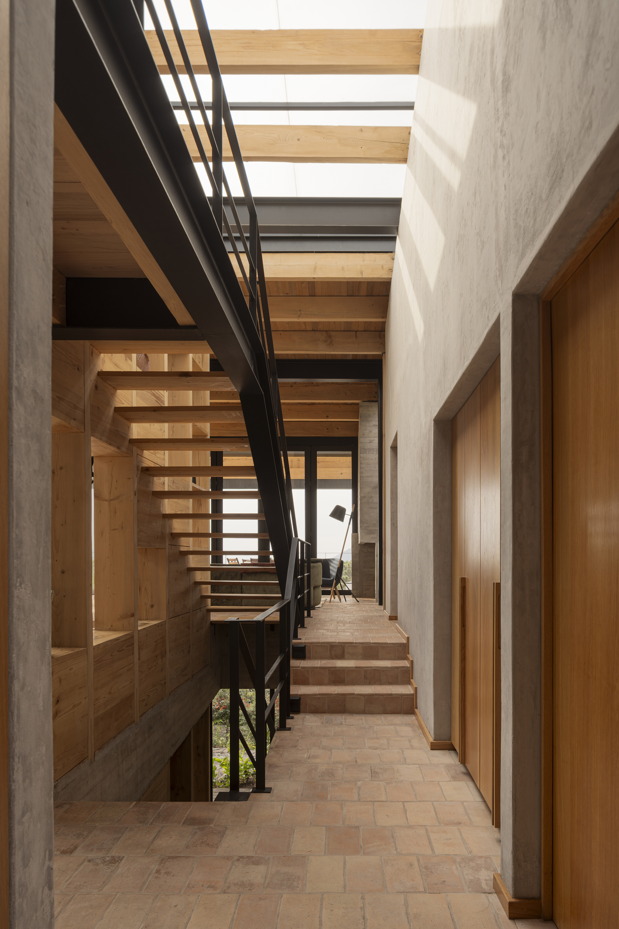 Corridor with open timber stair beside concrete walls and clay tile flooring under natural skylighting