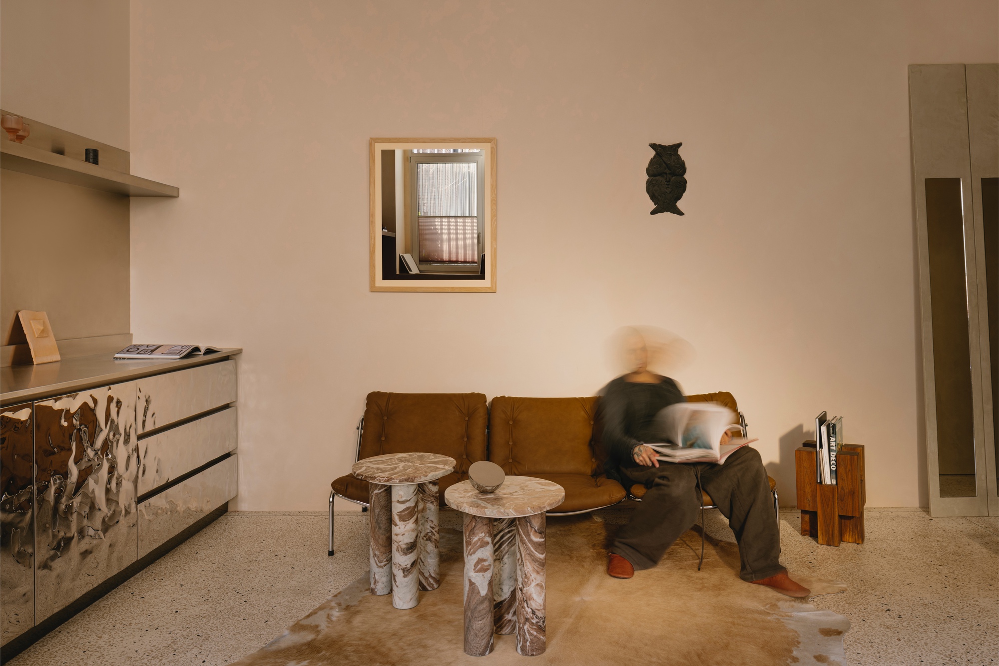 Living room with terrazzo flooring, tan sofa, carved timber side tables, and a person in motion
