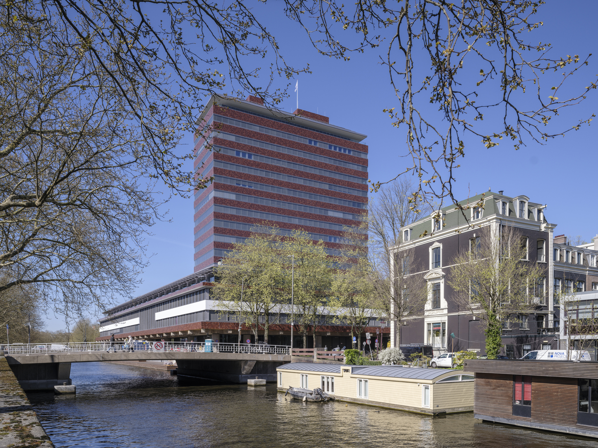 Horizontal brick and glass tower rising above a canal with houseboats and spring tree branches