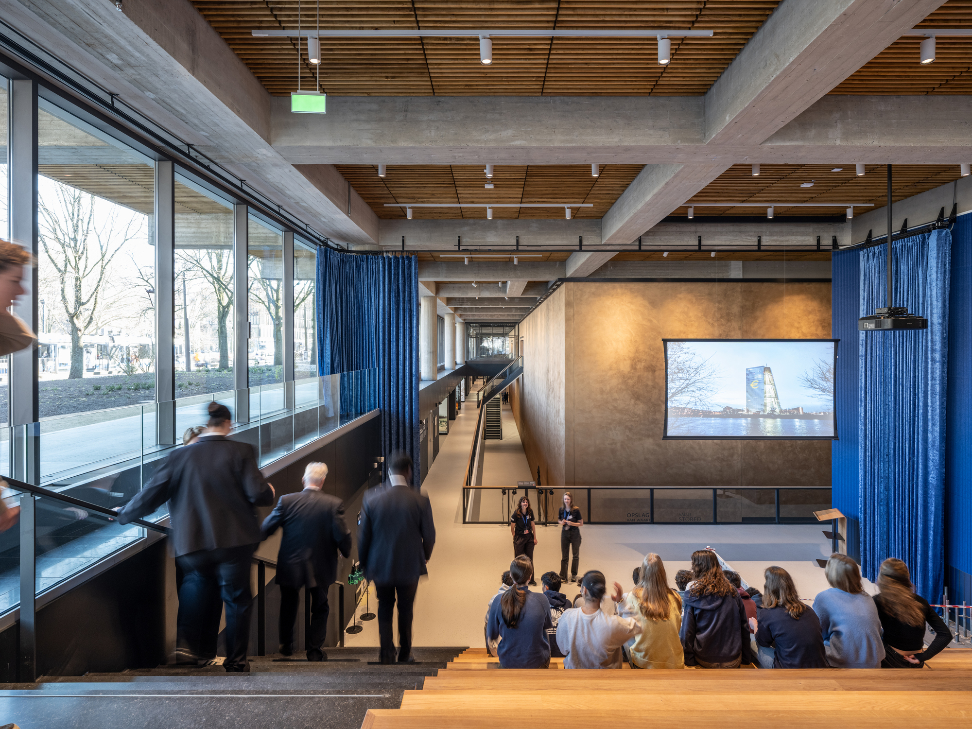 Interior auditorium with tiered wooden seating and concrete ceiling beams filled with an audience watching a presentation