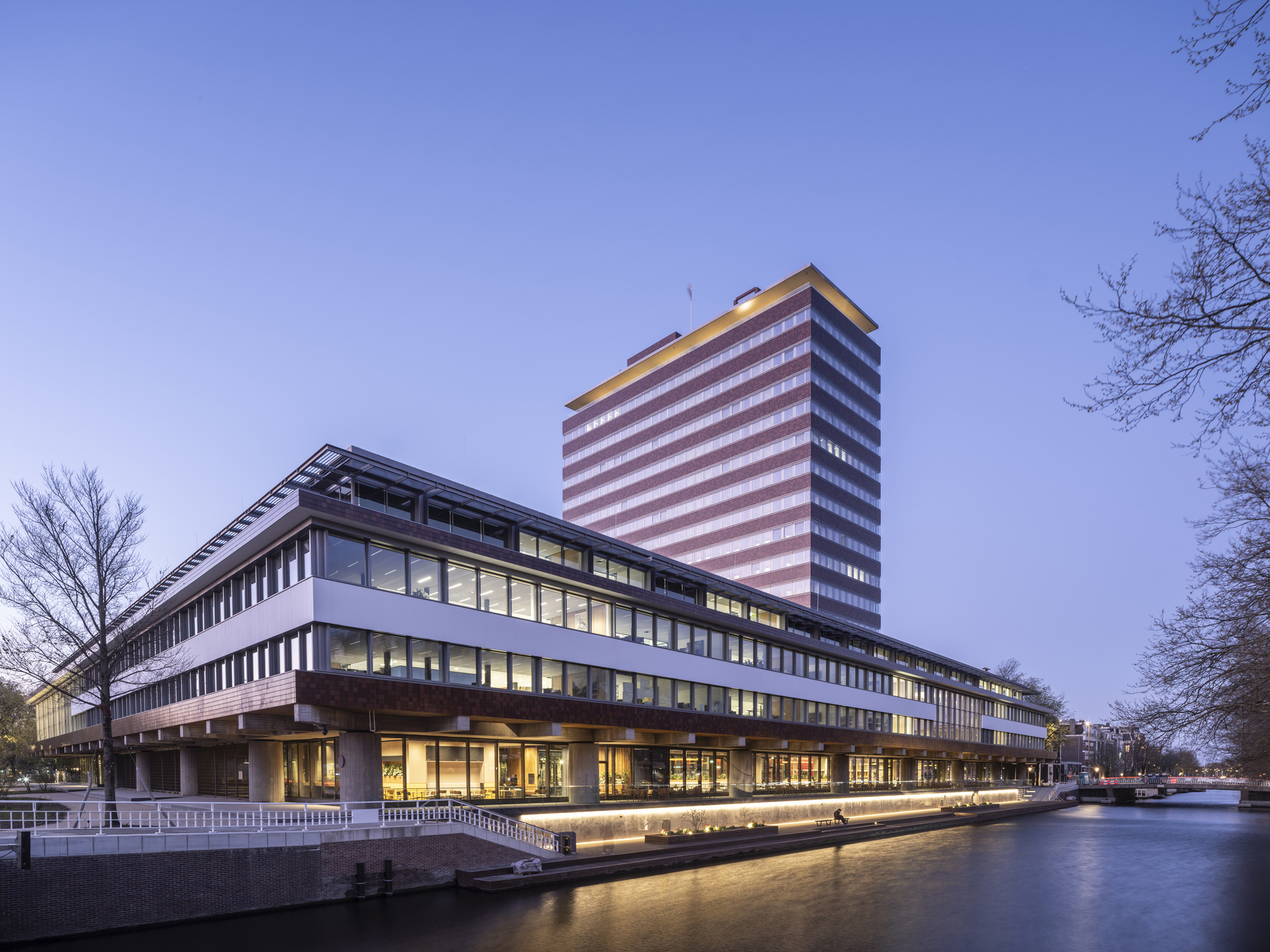 Canalside view of the horizontal building and illuminated tower at dusk with bare winter trees