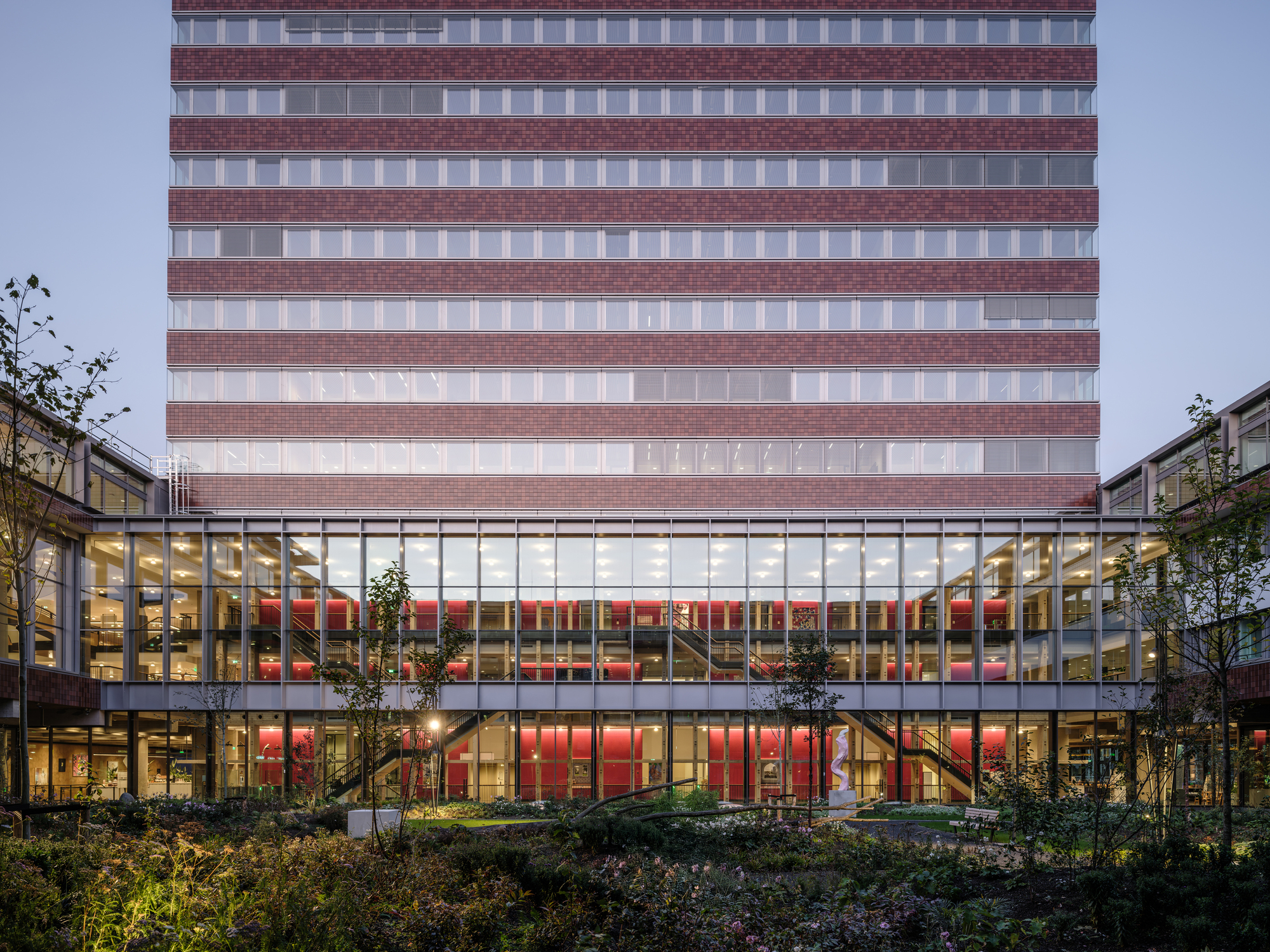 Illuminated glass and brick facade at dusk with tower rising above a planted courtyard