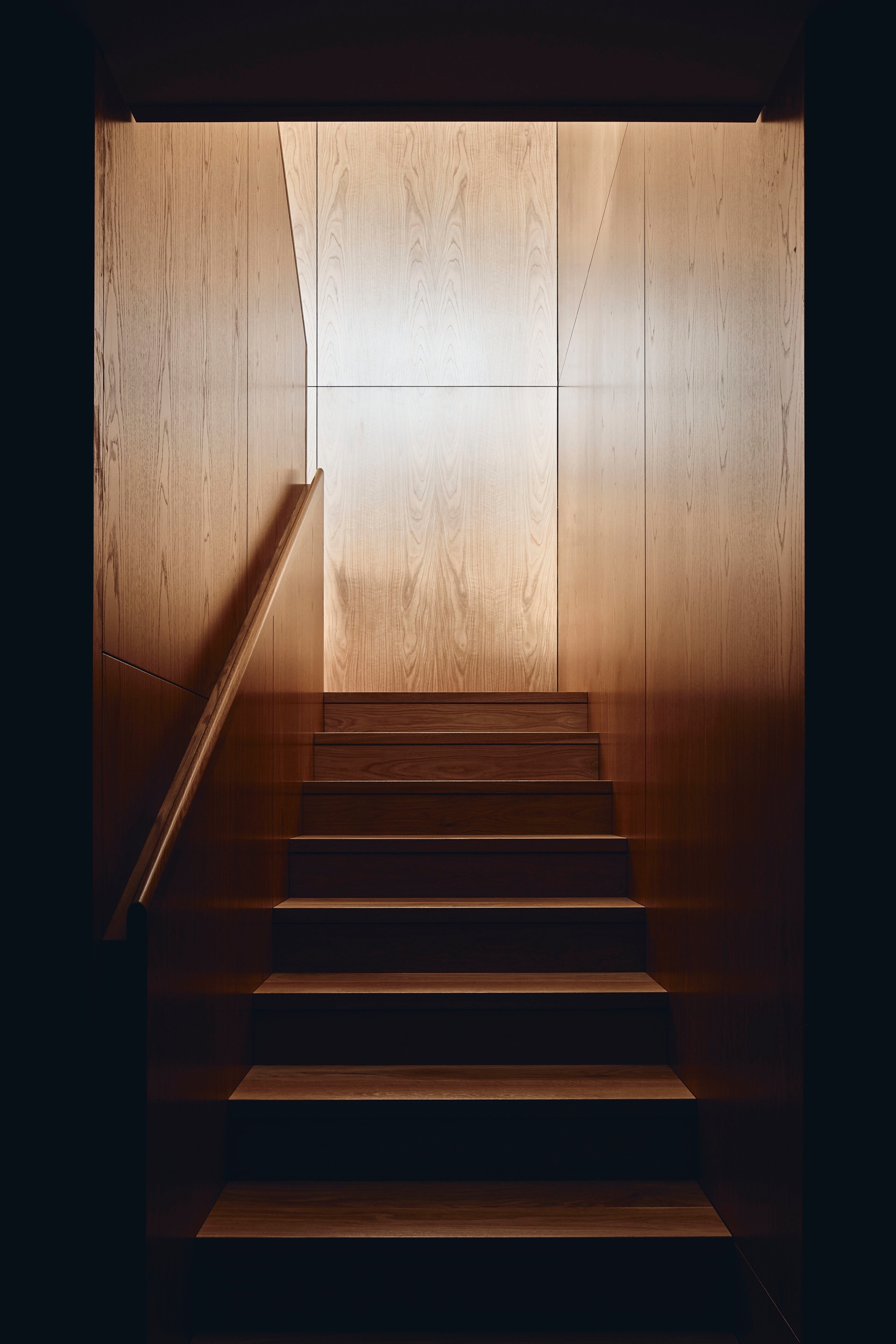 Timber staircase with cantilevered treads and blonde wood wall panels illuminated from above