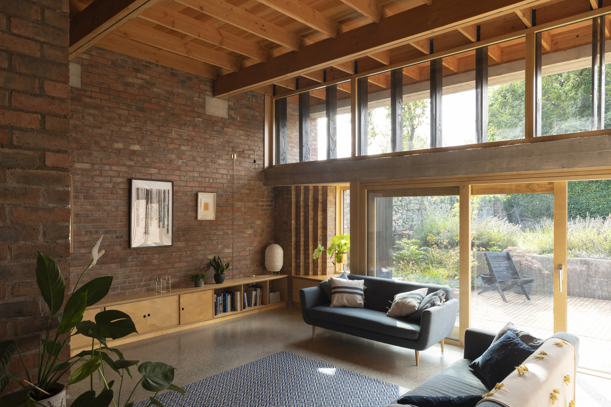 Double-height living space with exposed timber beams, brick walls and clerestory windows overlooking a terrace