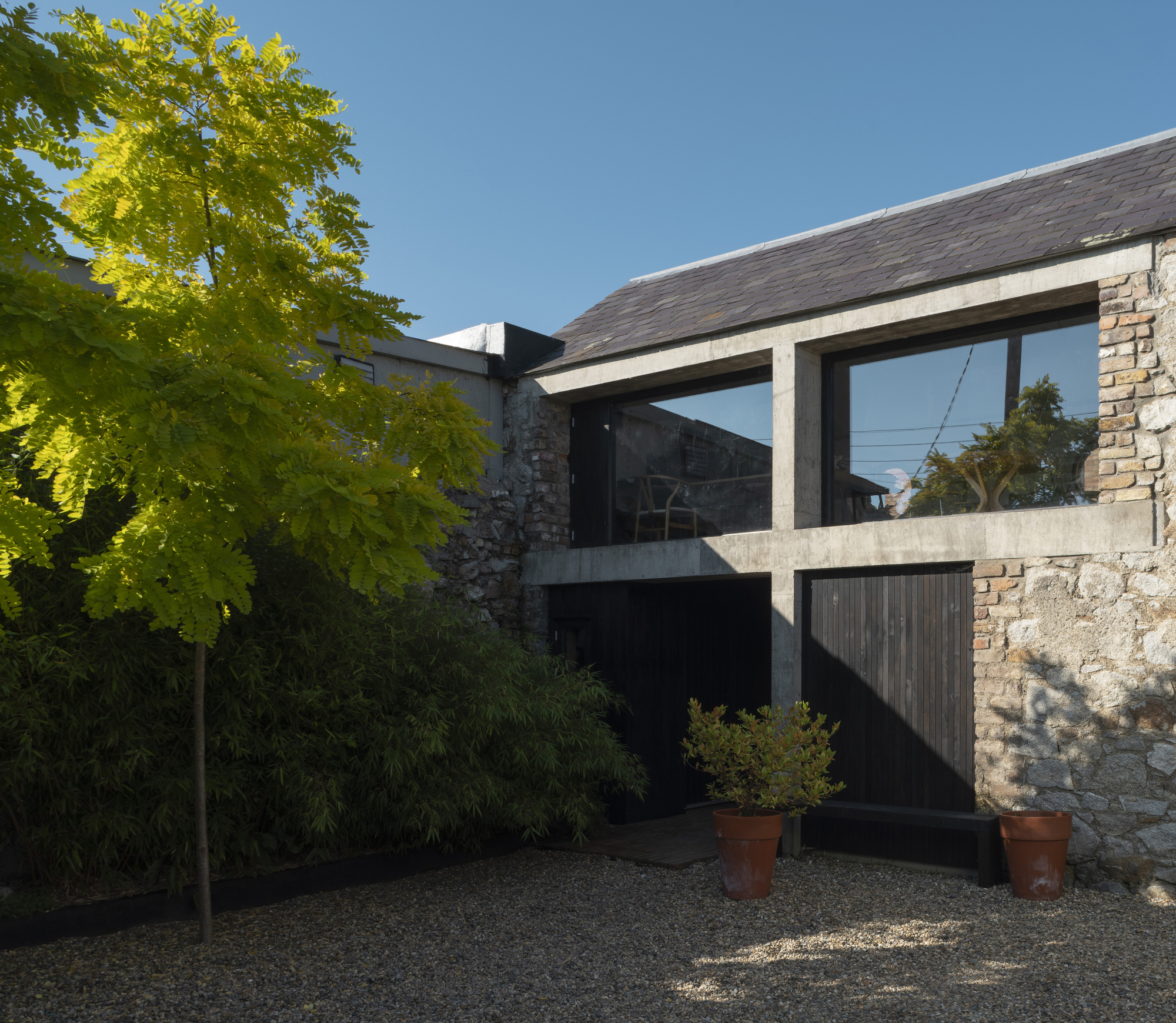 Slate-roofed extension with concrete frame and timber cladding beside a stone wall and potted trees