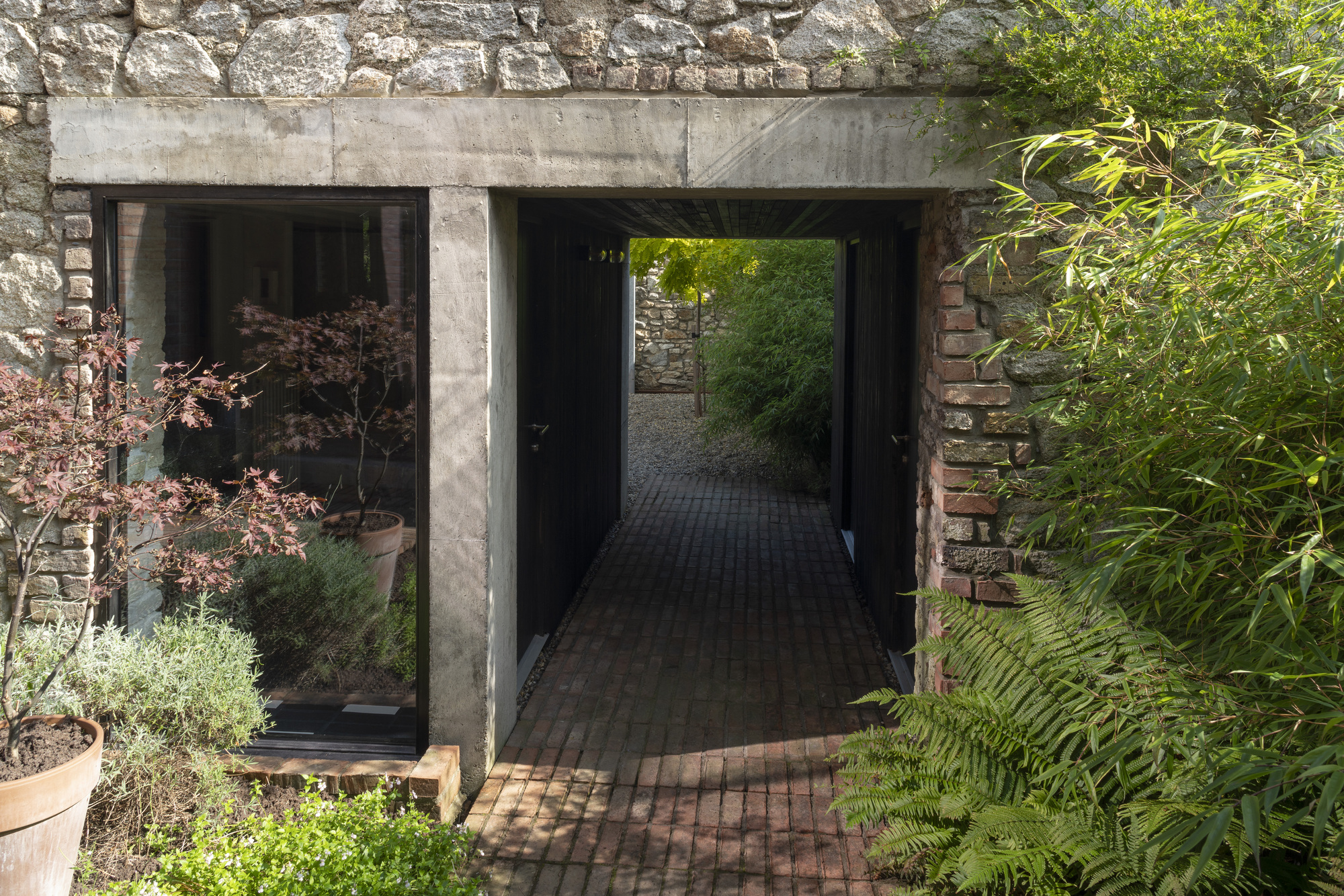 Concrete portal framing a brick-paved passage through a stone wall with planted beds and ferns