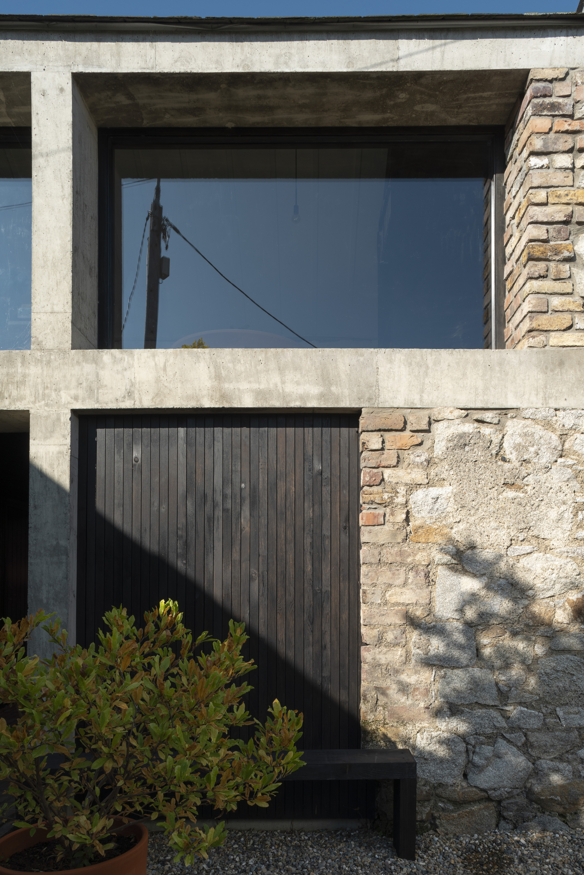 Layered facade showing concrete frame, black timber cladding, weathered stone wall, and planted shrubs in shadow