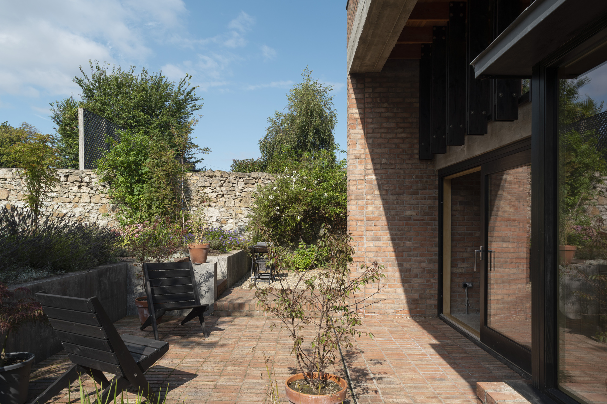 Brick paved terrace with potted trees and black timber seating against a stone boundary wall