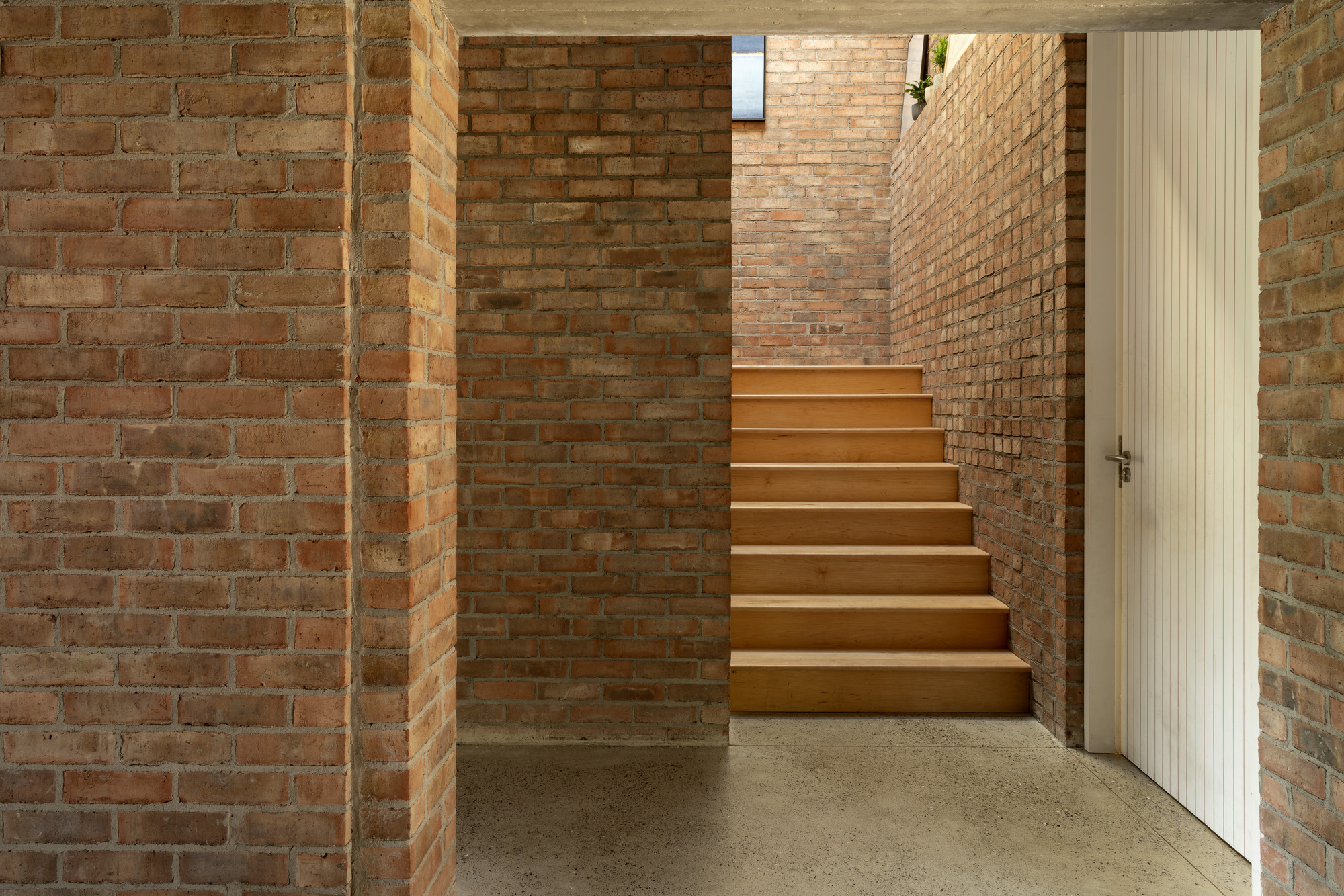 Timber staircase rising between exposed brick walls with a white ribbed door and polished concrete floor