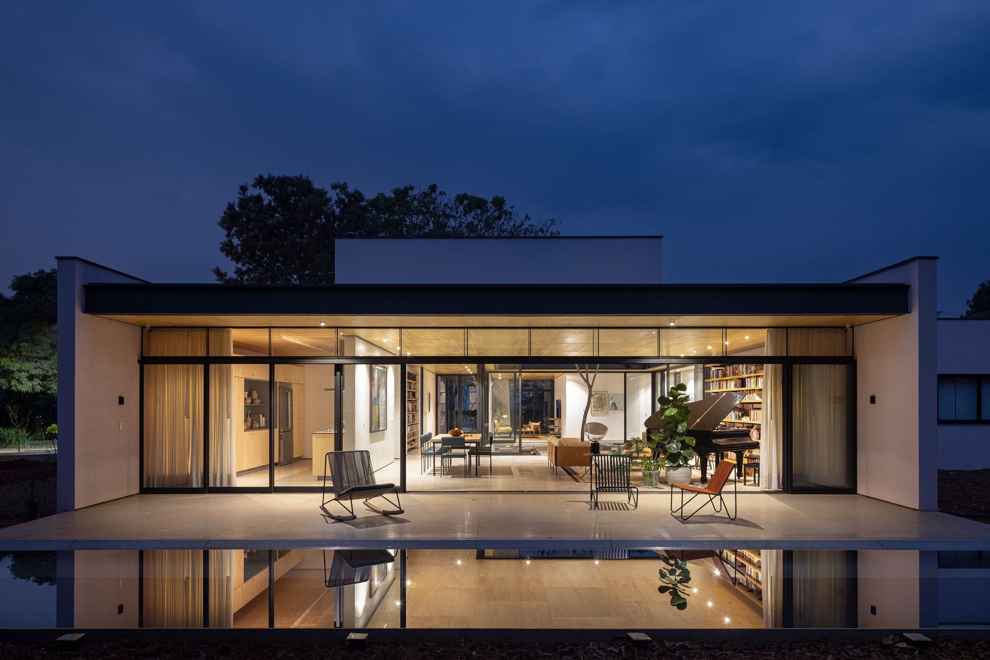 Fully glazed facade opening to terrace and reflecting pool with interior furniture visible at twilight