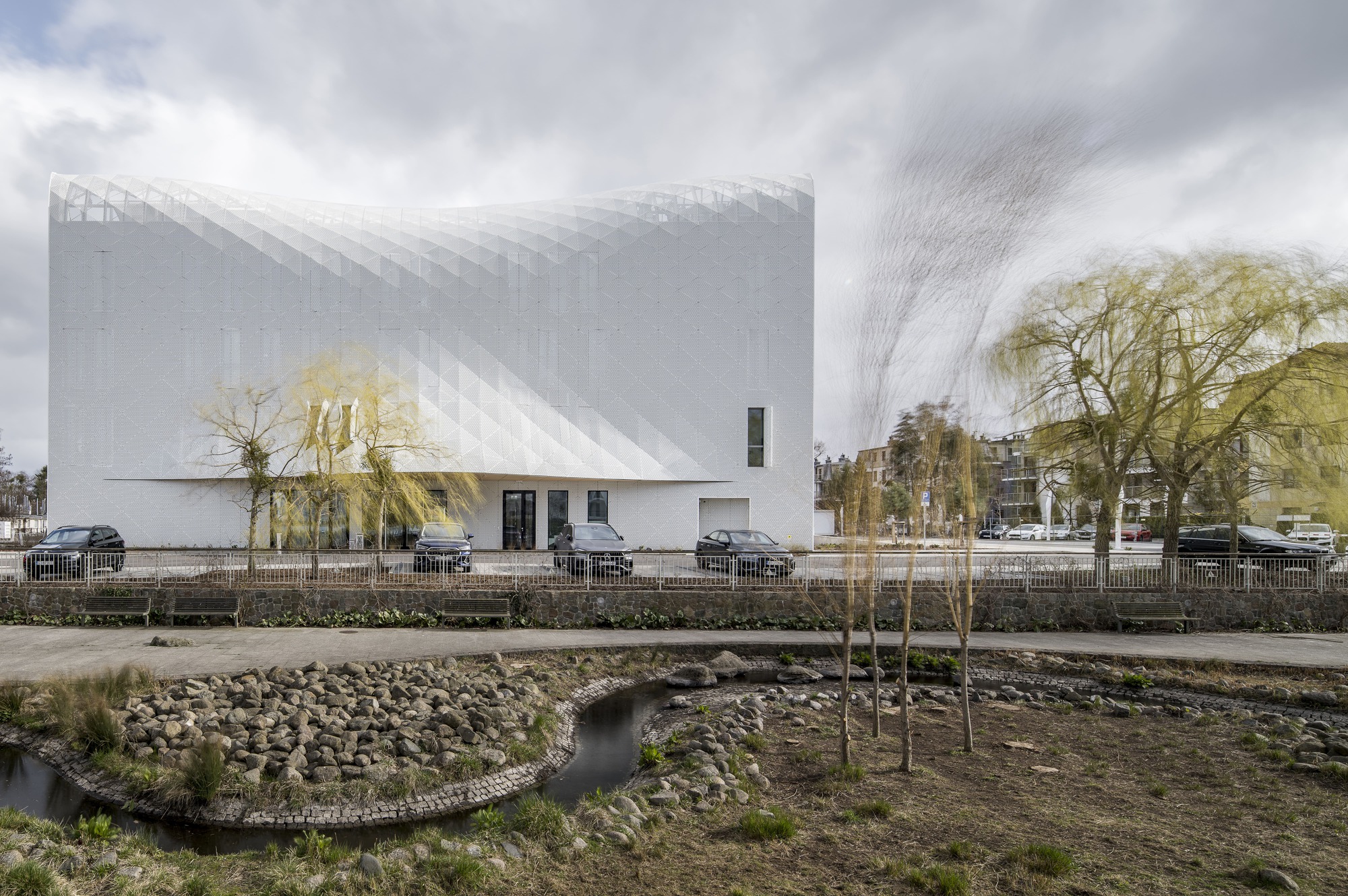 Street view of the textured white facade and curved volume across from a landscaped stream with young trees