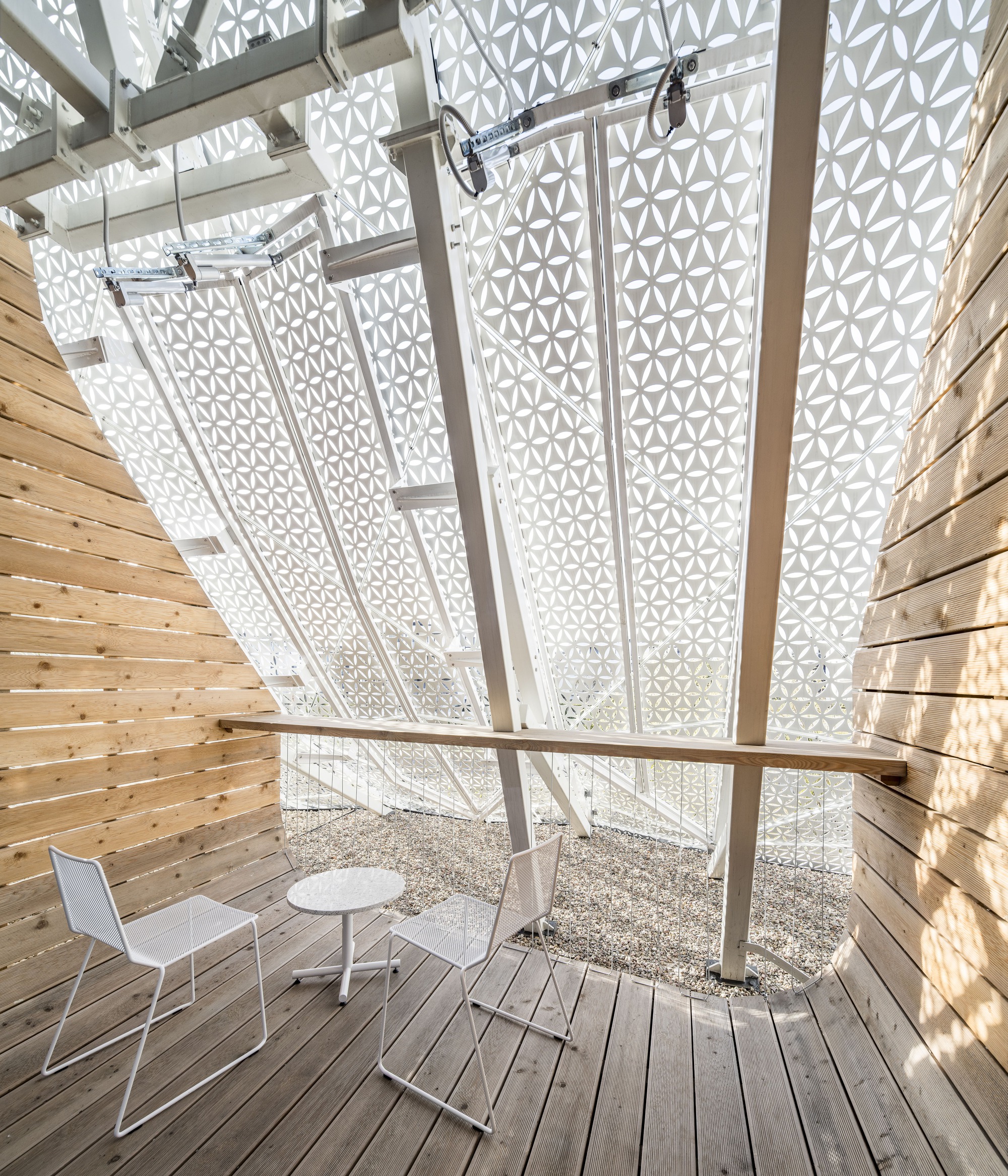 Timber deck terrace enclosed by perforated white panels with dappled sunlight and furniture