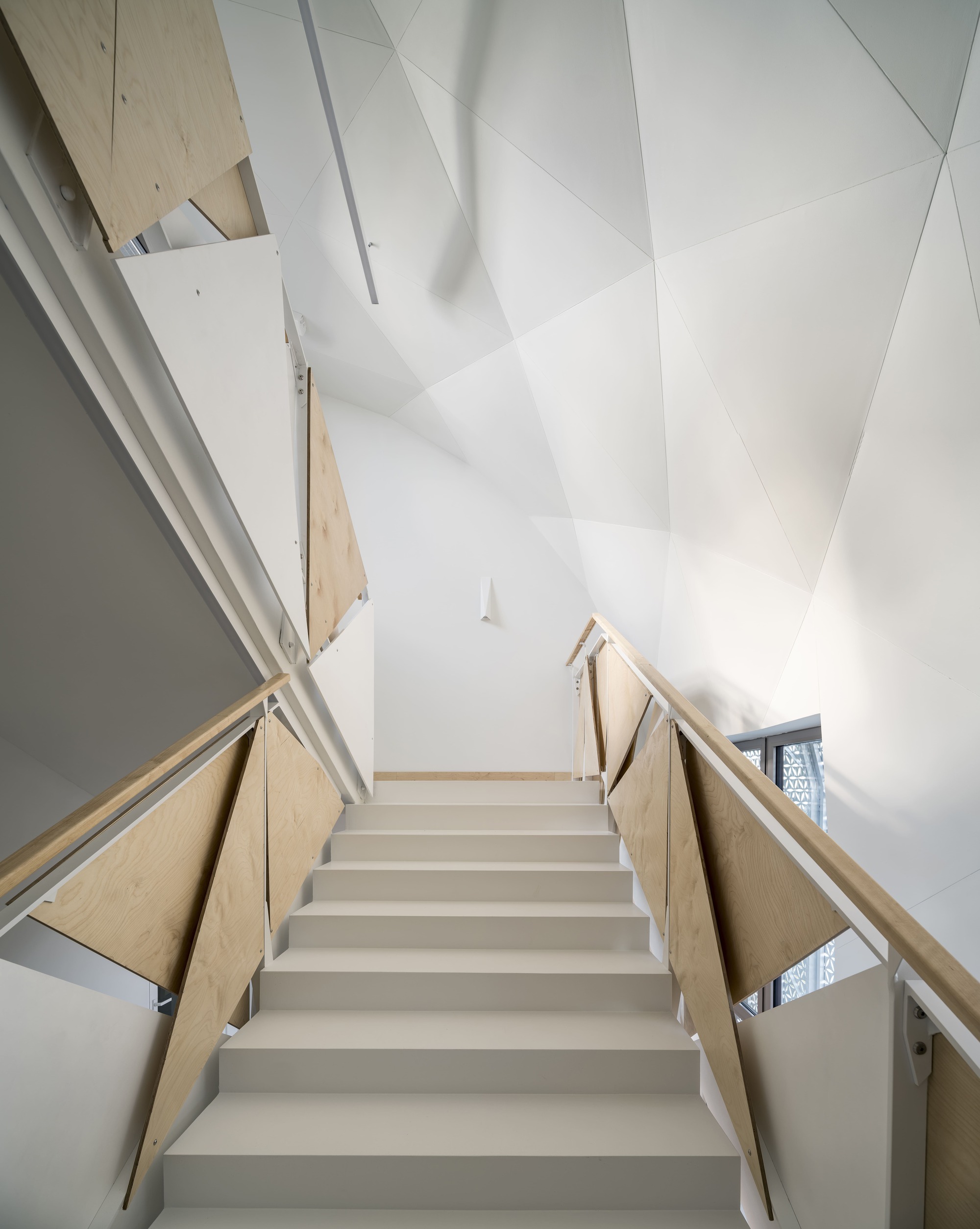 Interior staircase with plywood balustrades and white faceted ceiling under natural light