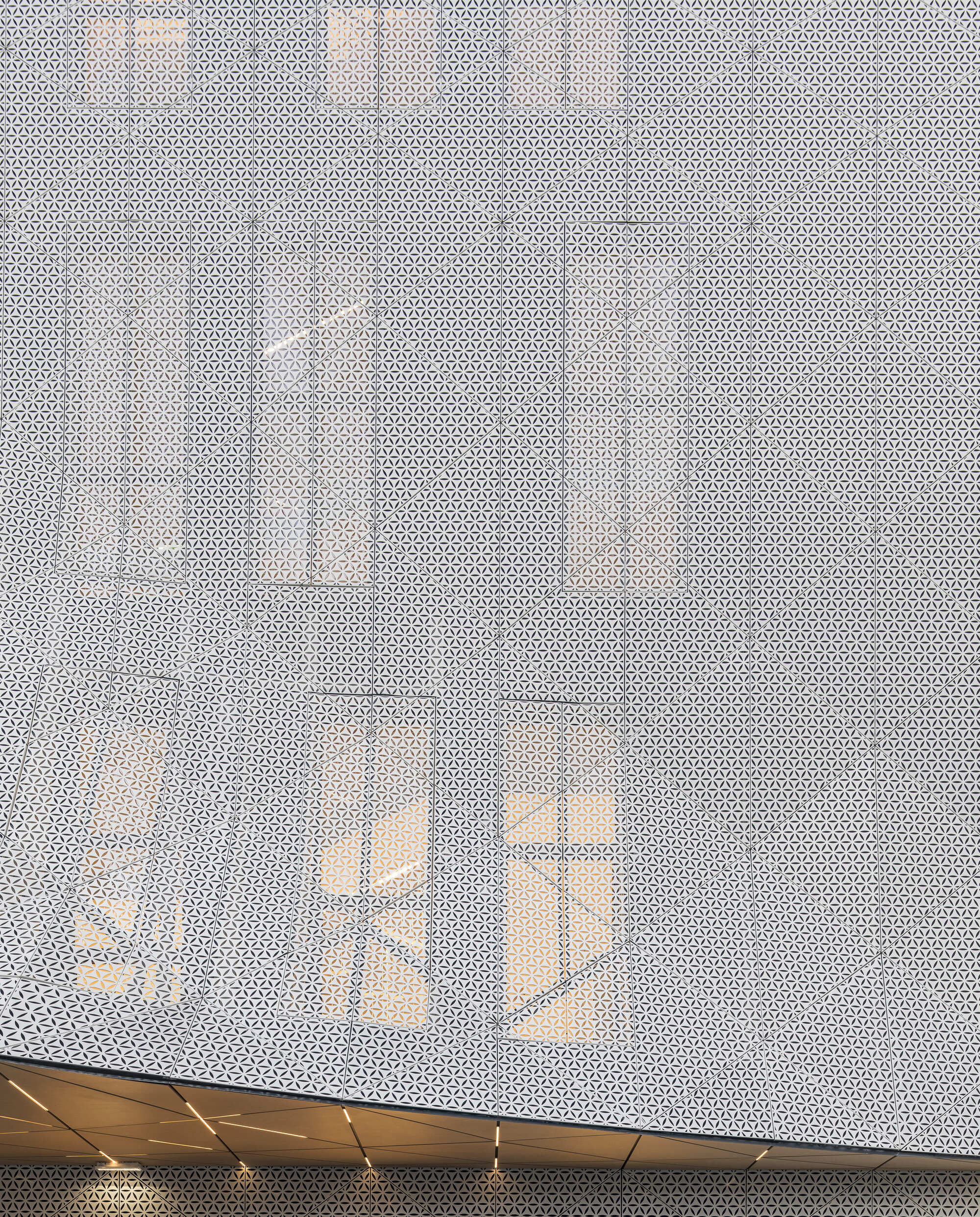 Perforated metal screen wall with timber beams visible through the translucent surface at dusk