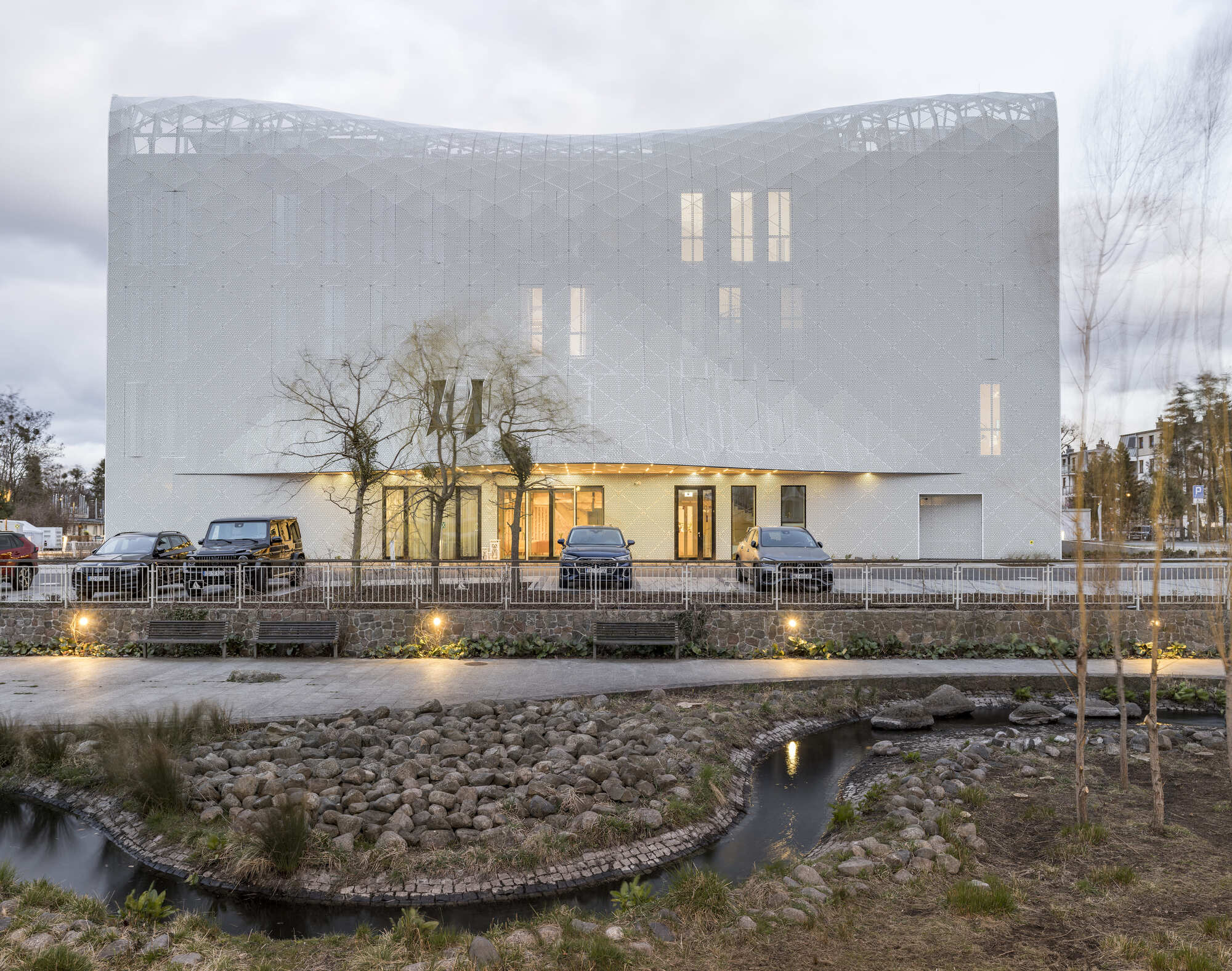 Curved perforated metal facade with illuminated base and reflecting pool at dusk