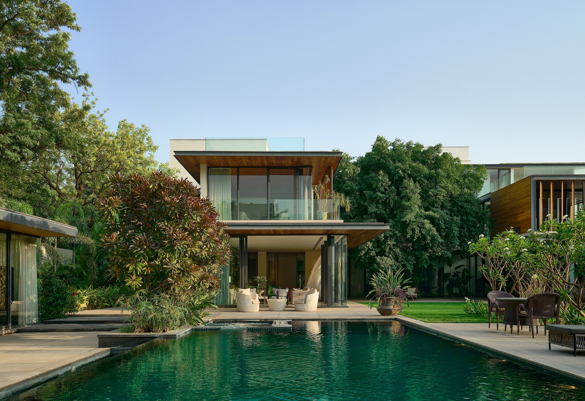Two-storey glass and timber residence with pool terrace framed by mature trees under afternoon light