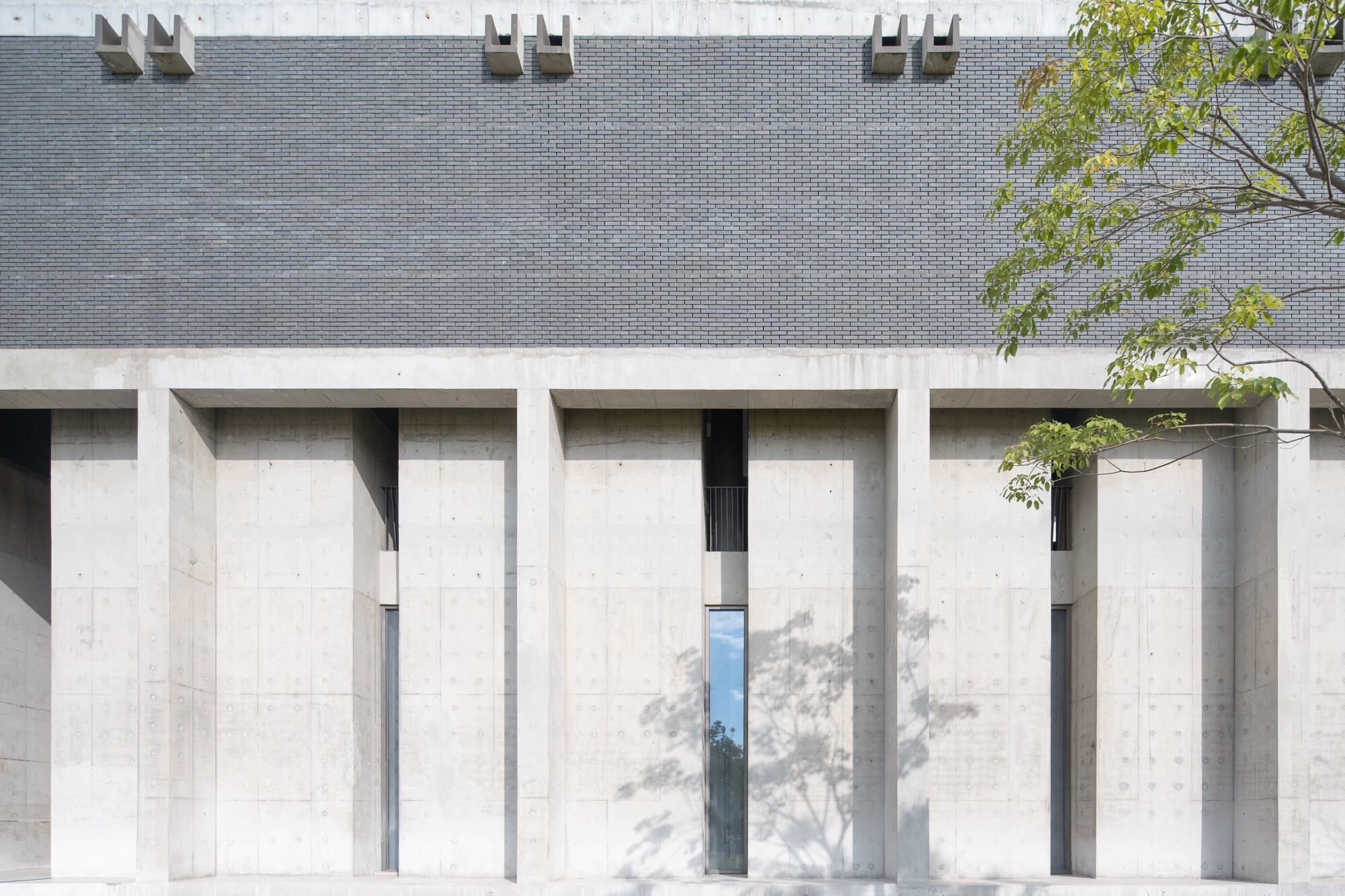 Facade with recessed concrete columns below grey brick wall and dappled tree shadows in daylight