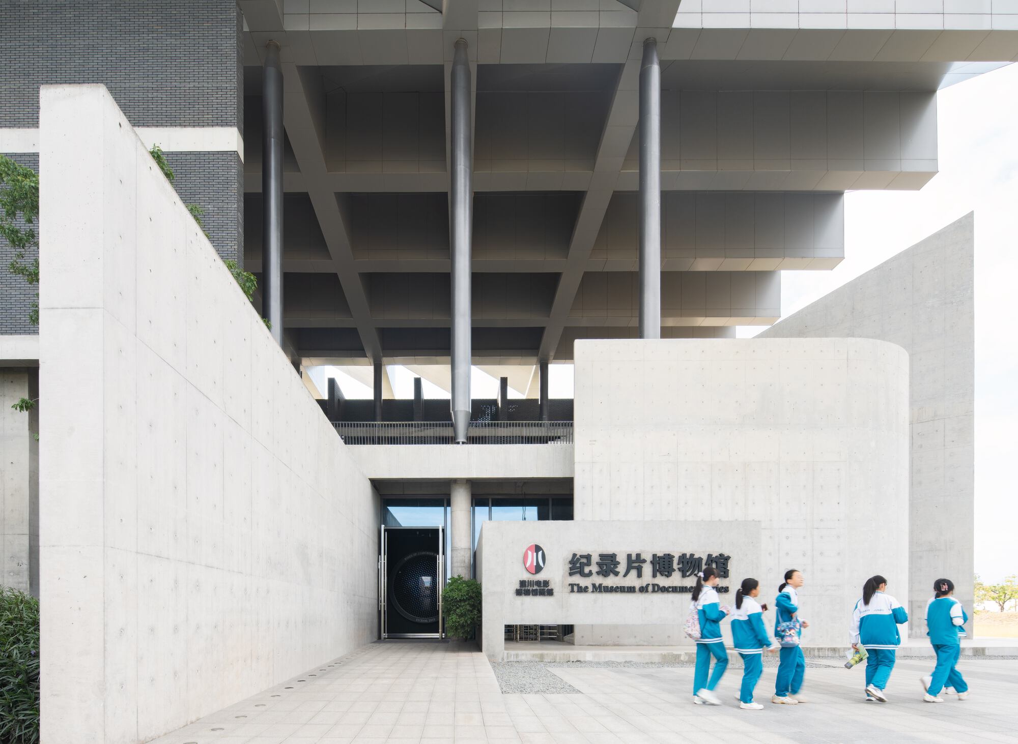 Courtyard entrance with cylindrical concrete element and group of uniformed visitors walking across the paving