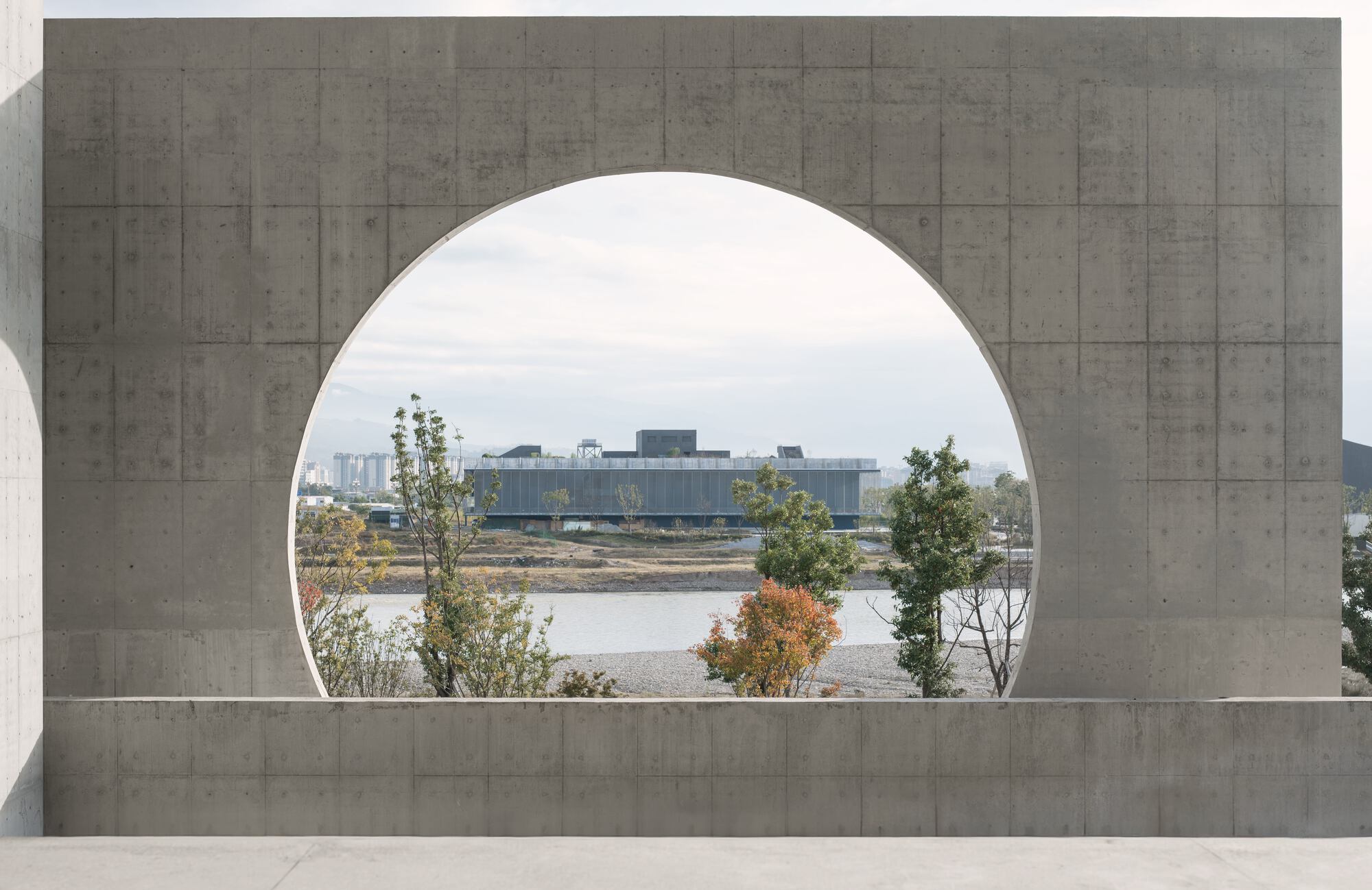 Large circular aperture in a board-formed concrete wall framing a view of the river and distant trees