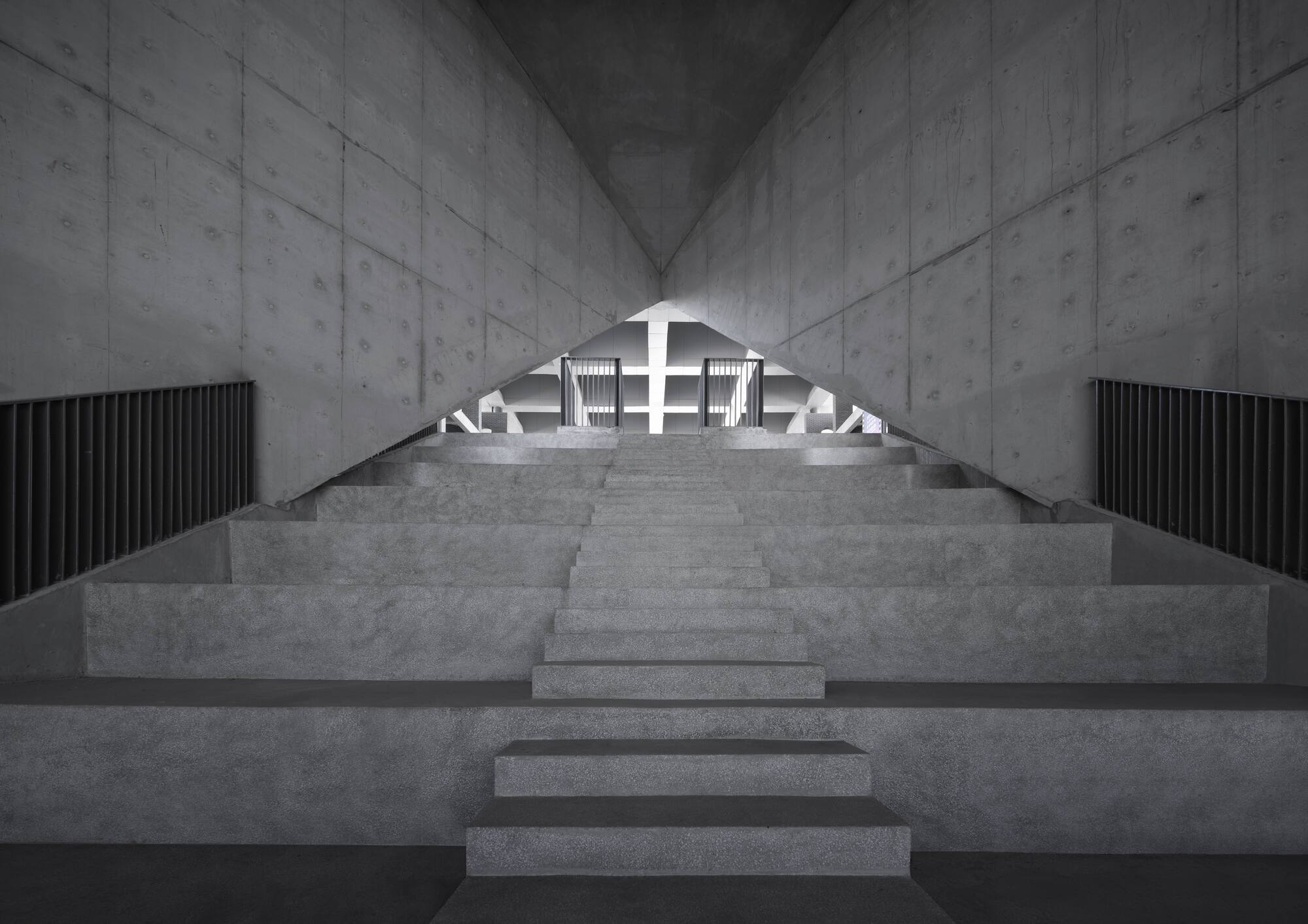 Central concrete staircase rising between board-formed walls toward a backlit arched opening