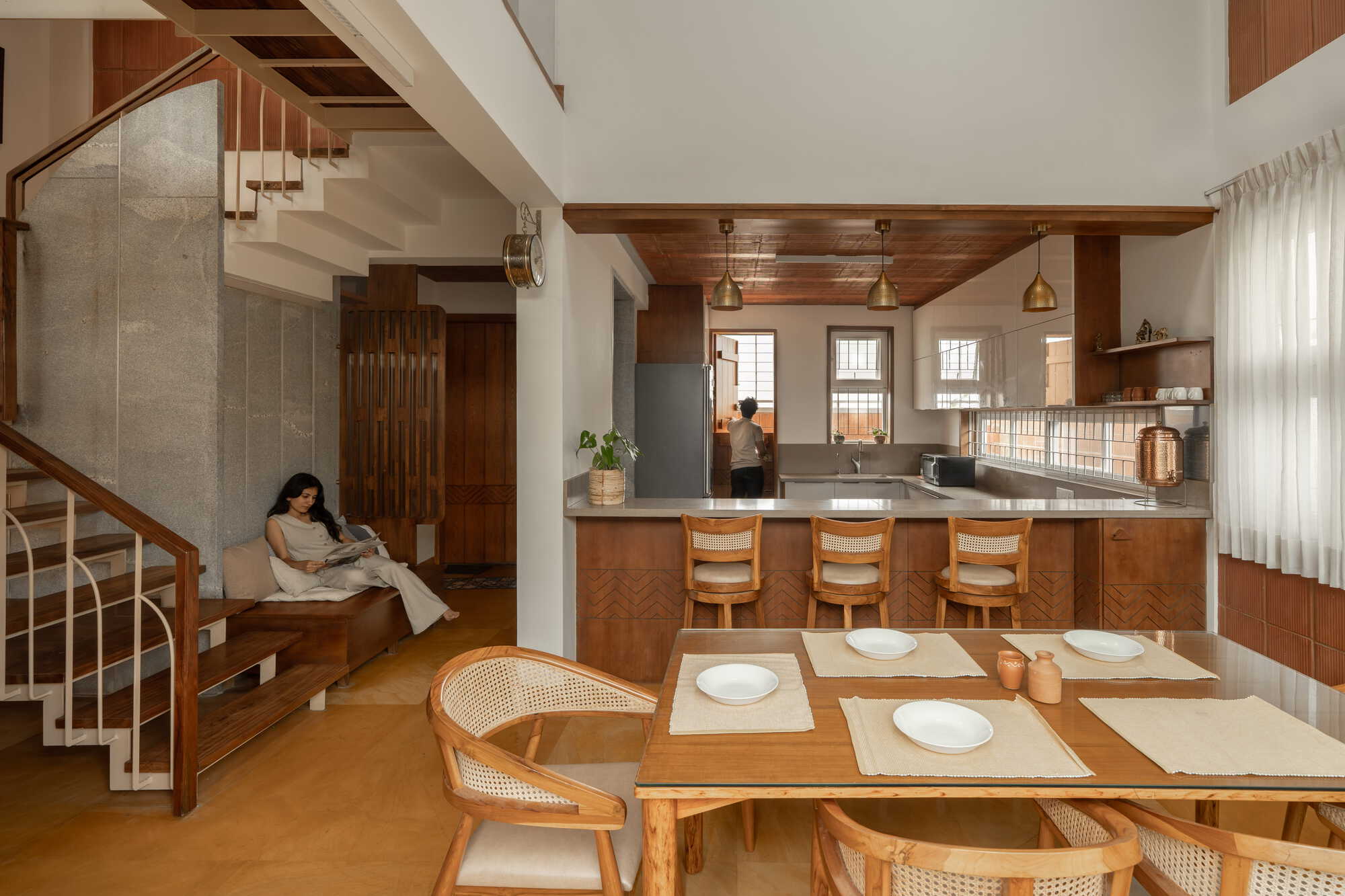 Open plan living space with terracotta tile walls, timber dining table and staircase rising through a double-height volume
