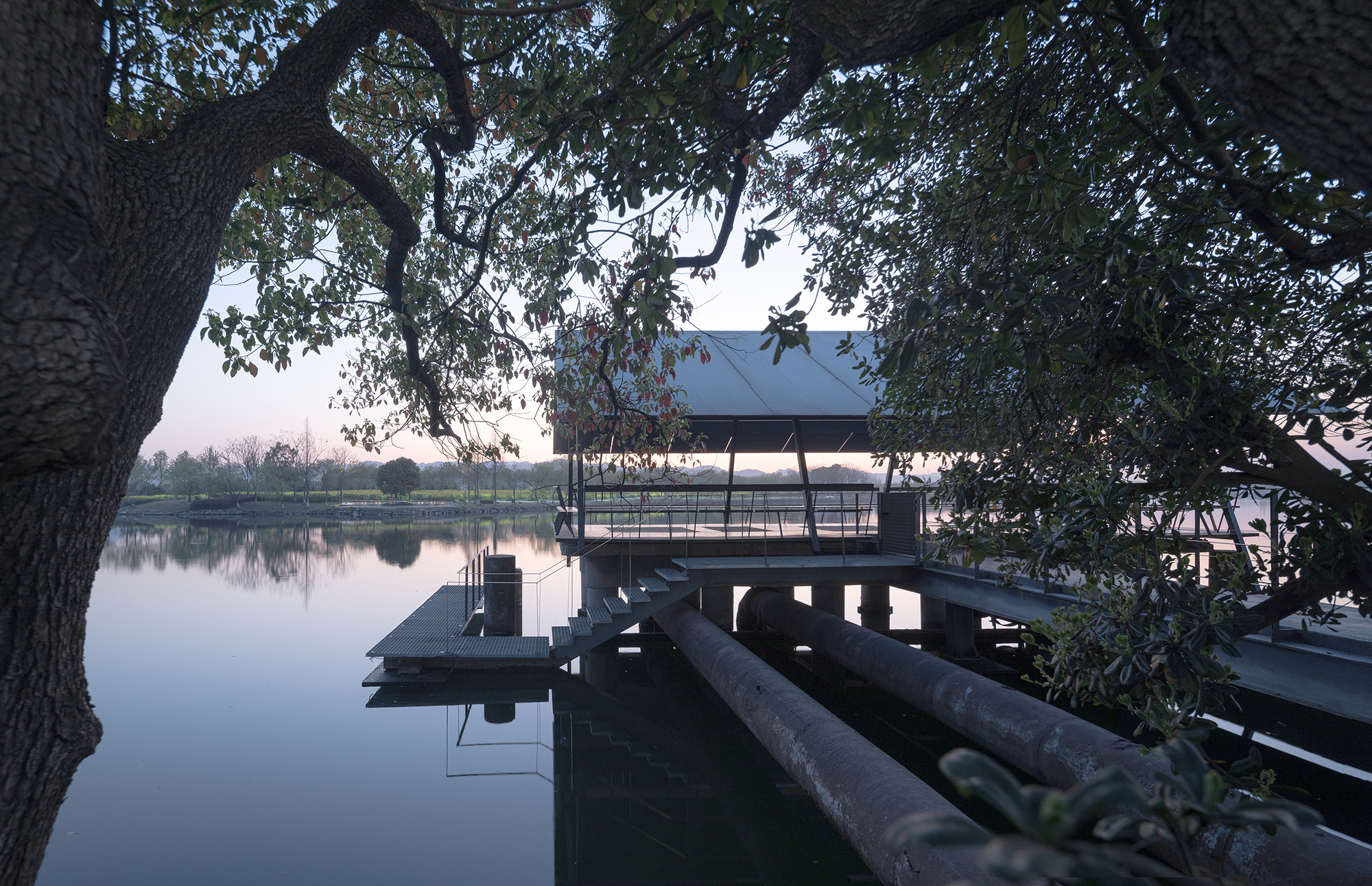 Elevated pavilion with metal roof and timber deck extending over still water at dusk beneath overhanging tree branches