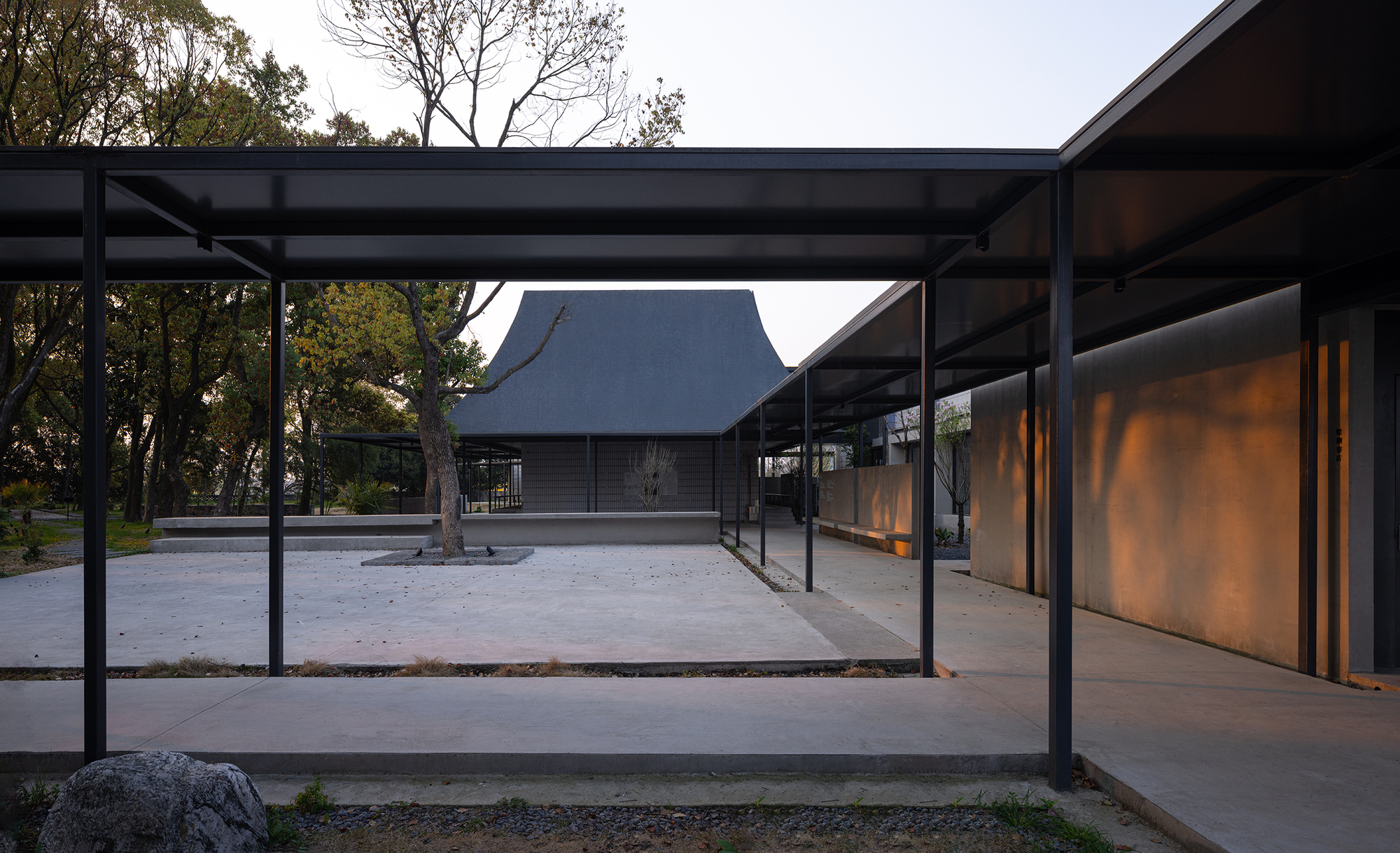 Covered walkway with steel frame and concrete floor connecting two pavilions with pitched roofs in early evening light