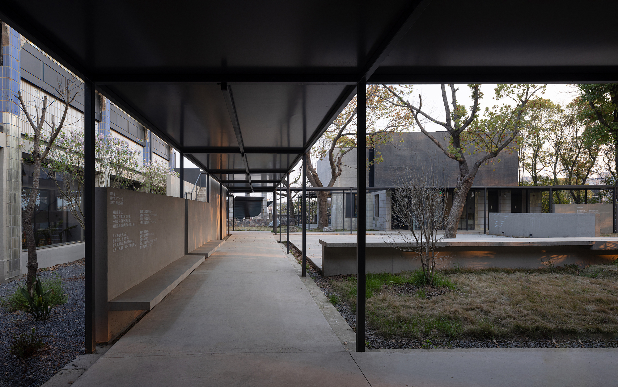 Corridor with steel columns and translucent roof panels opening onto courtyard with planted gravel and scattered trees
