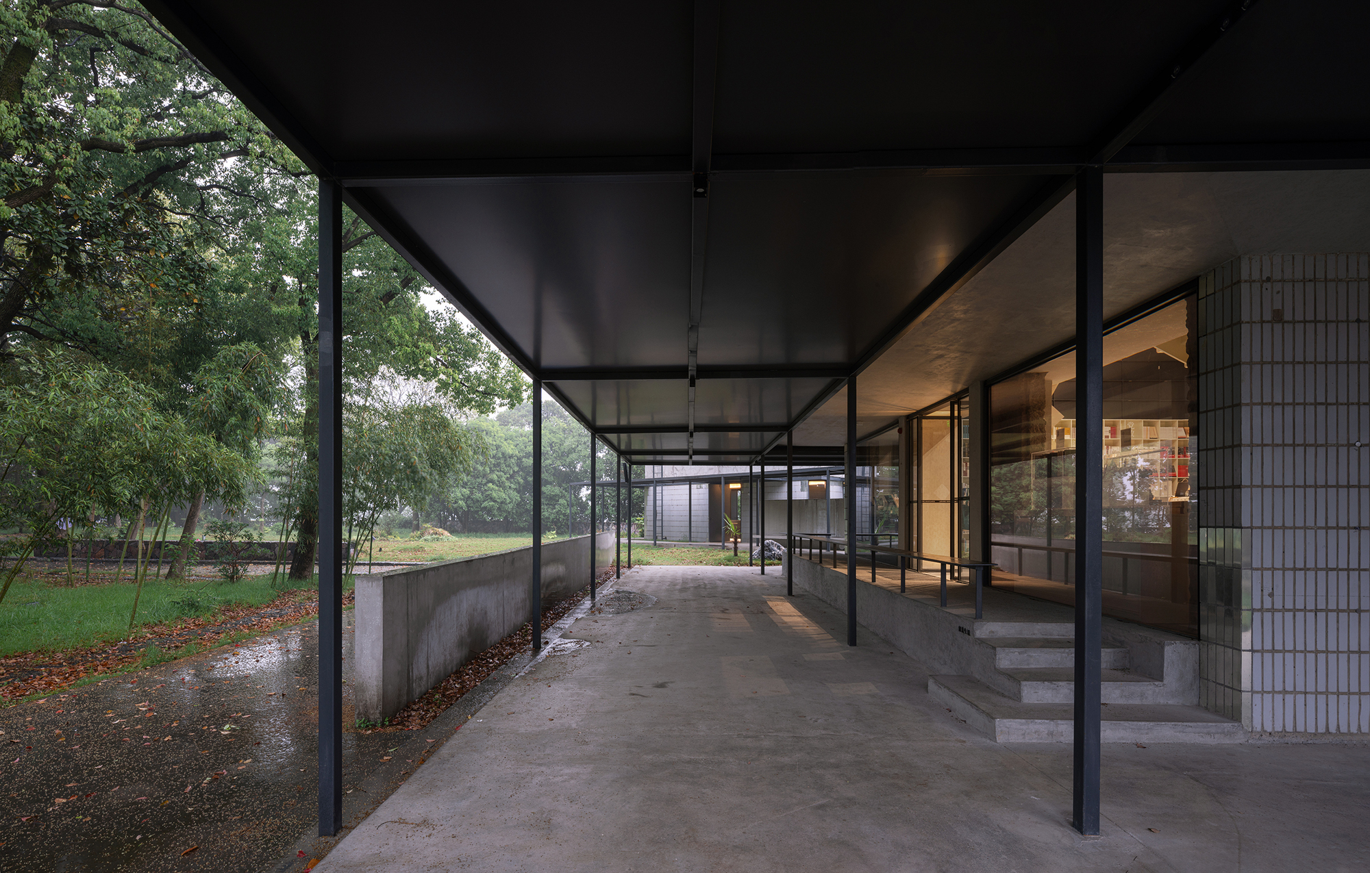 Covered entry walkway with slender steel columns and full-height glazing overlooking a park in wet weather