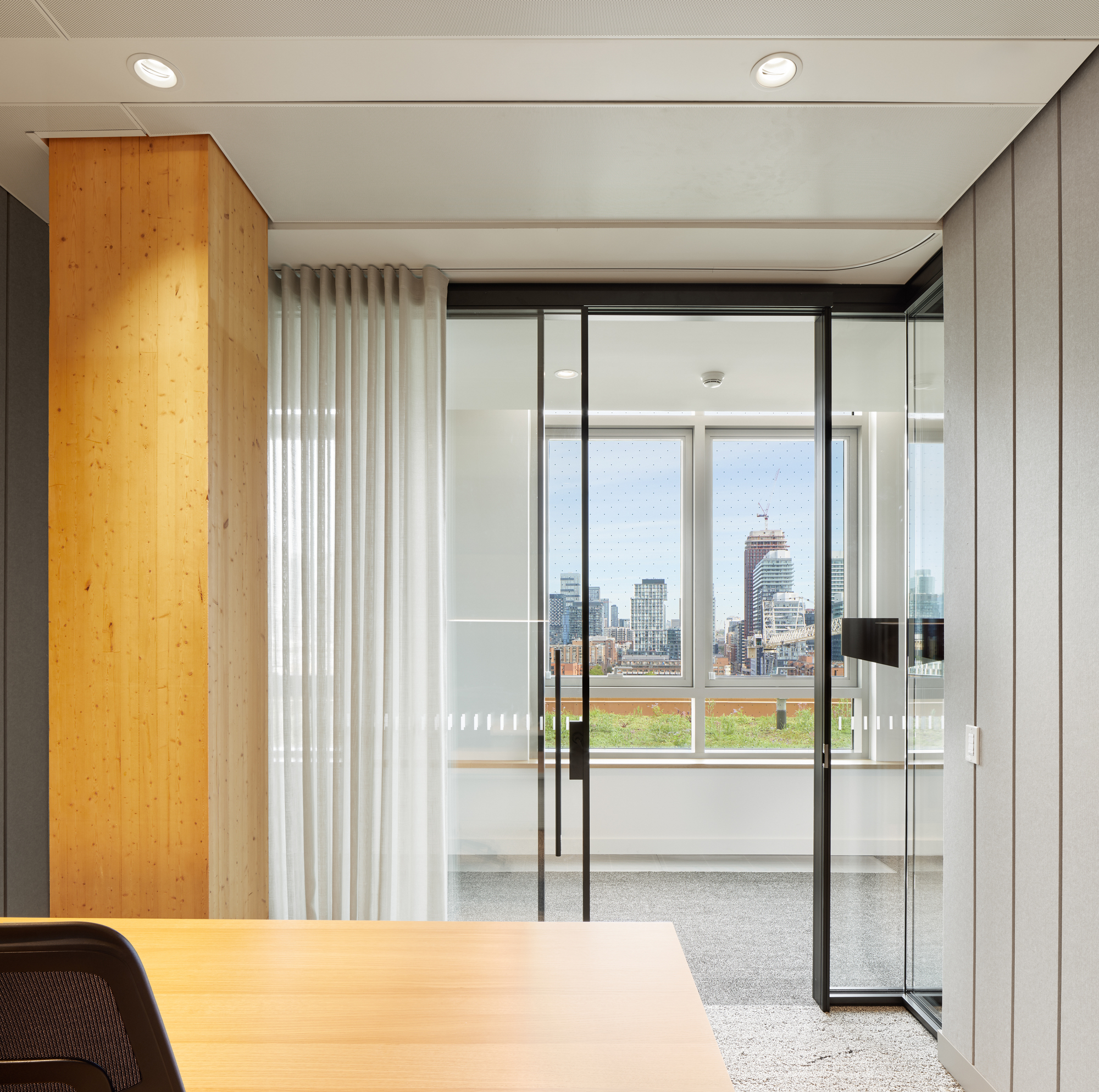 Glass partition with white curtain framing a window view of the urban skyline and rooftop plantings